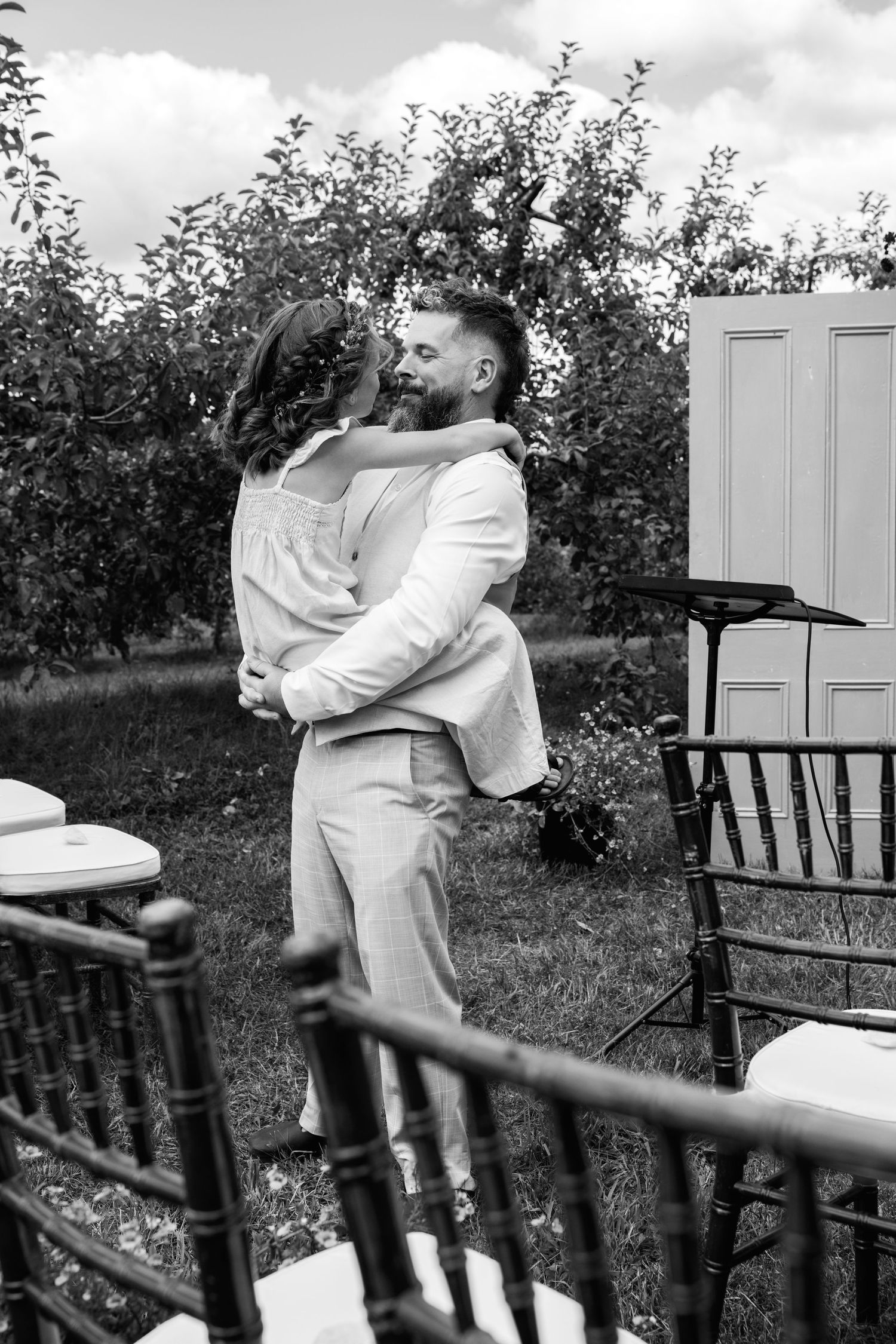 Groom lifts bride in his arms during outdoor wedding ceremony in apple orchard, black and white photo.