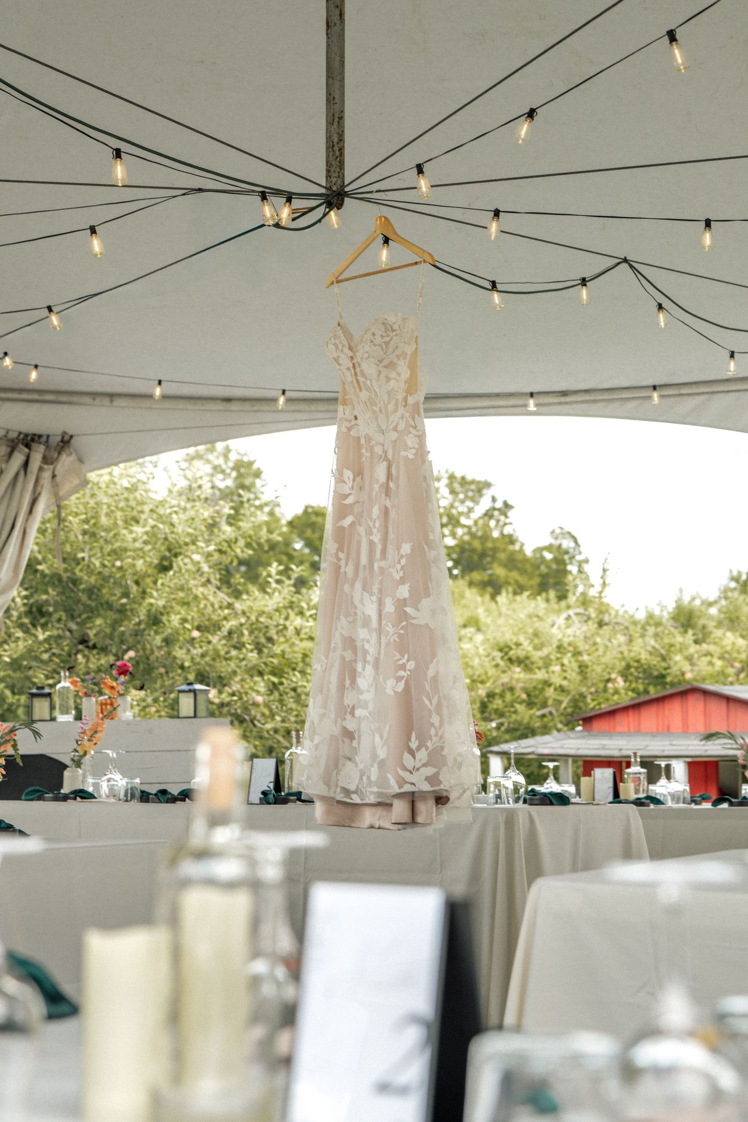 Elegant lace wedding dress hanging from a wooden hanger under a tent with string lights.