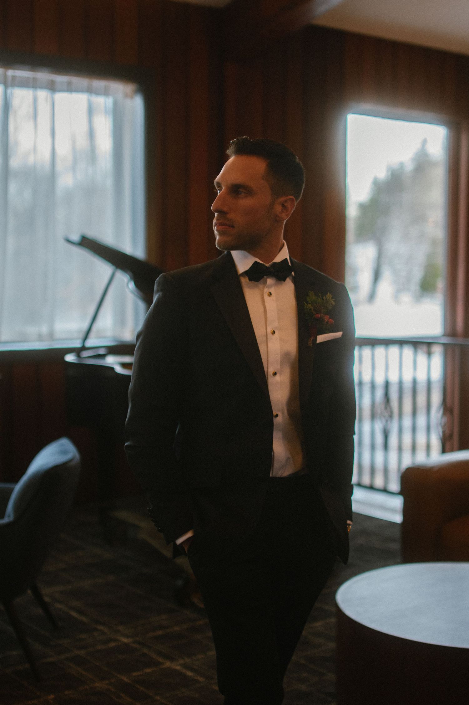 Groom in black tuxedo with bow tie and boutonniere standing in elegant wood-paneled room with snowy windows.