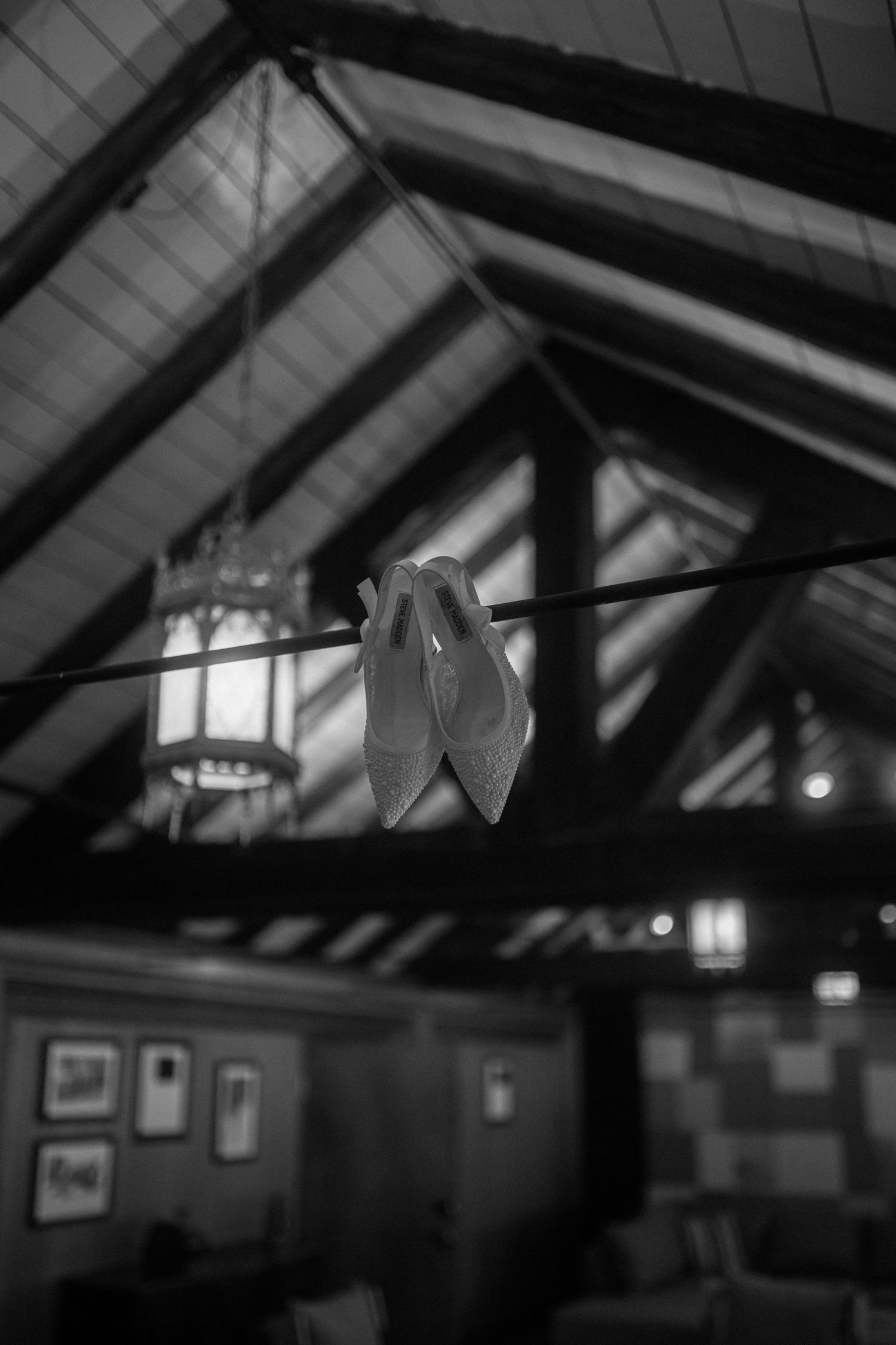 Elegant bridal heels hanging from rustic wooden ceiling beams in a cozy lodge, captured in black and white.
