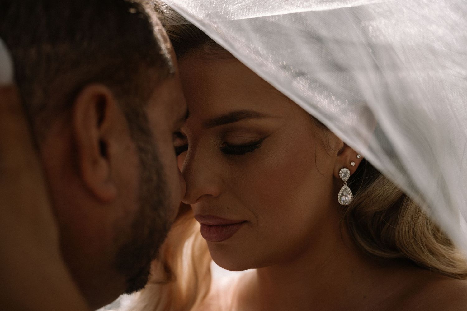 Intimate close-up of bride and groom nearly kissing beneath a flowing white wedding veil.