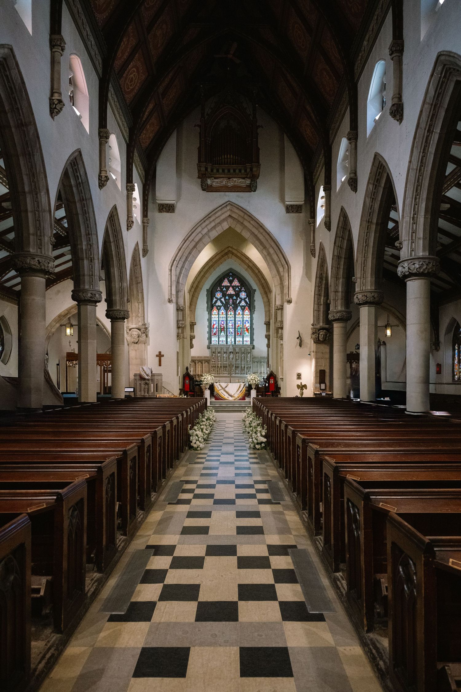 Gothic church interior with wooden pews, checkered floor, and stained glass altar decorated with white wedding flowers.