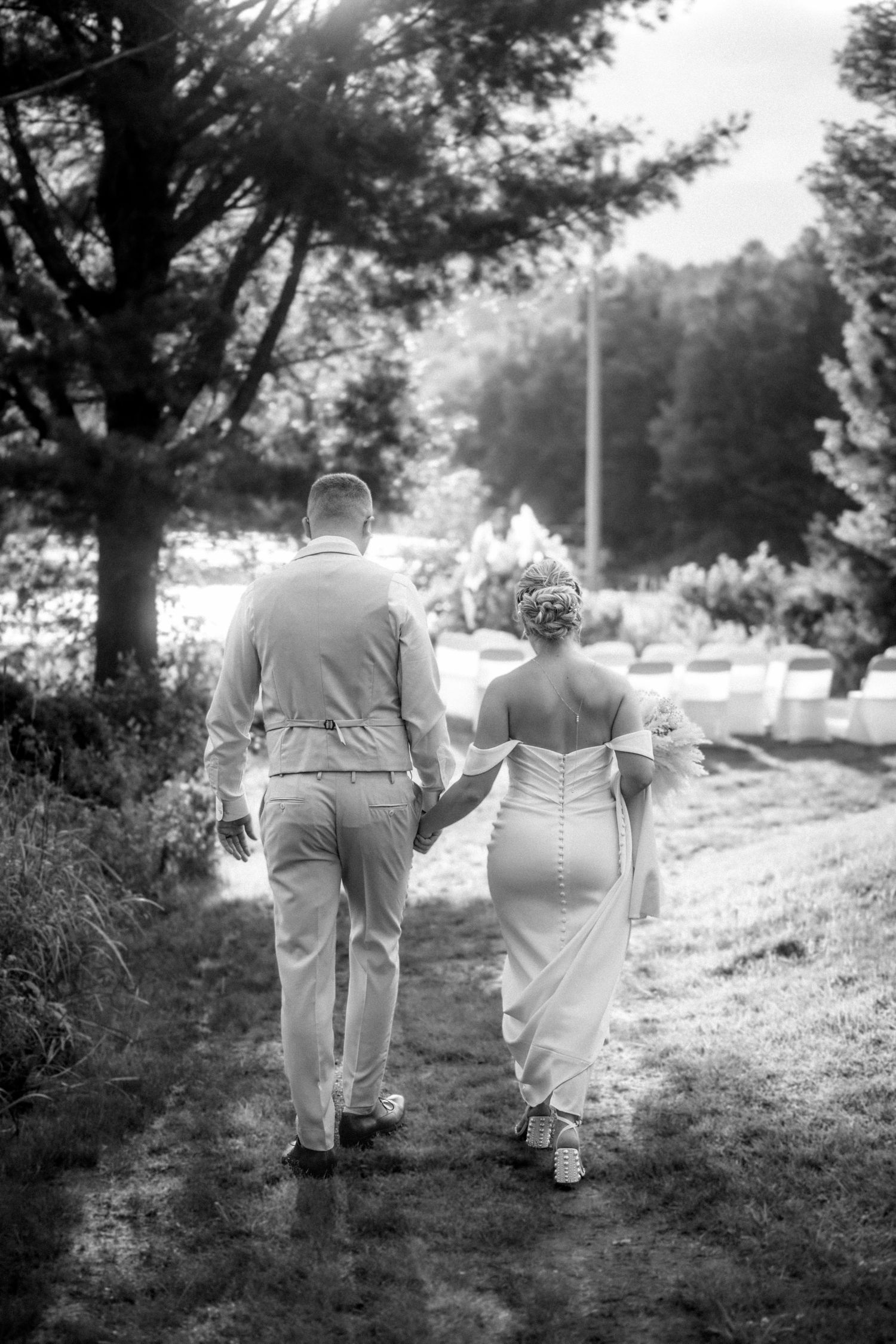 Black and white photo of bride and groom walking hand in hand through a garden path at their outdoor wedding.