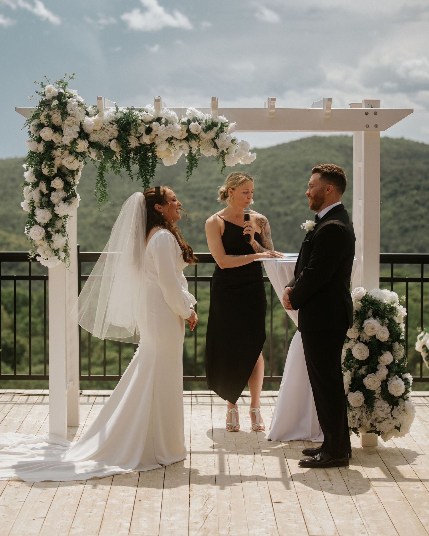 Bride and groom exchange vows under a white floral arch on an outdoor terrace with mountain views.