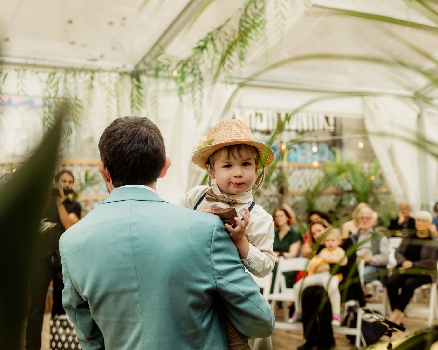 Man in teal suit holds smiling toddler in hat at elegant indoor wedding ceremony with guests seated.