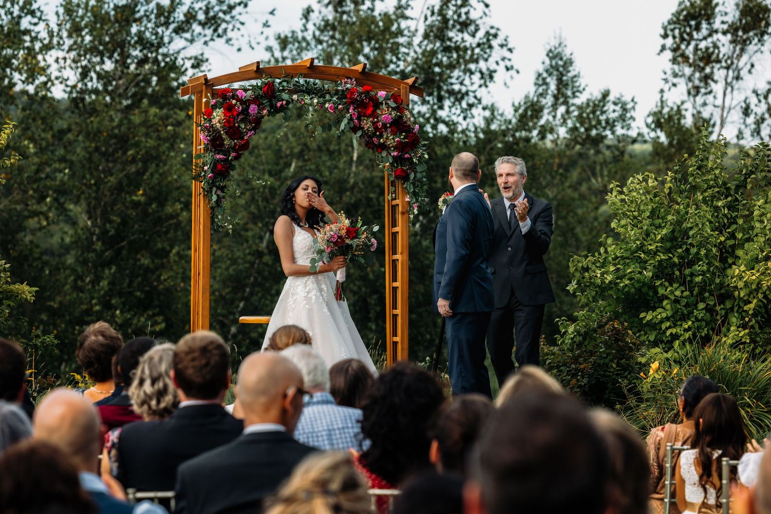 Outdoor wedding ceremony with bride and groom at floral arch, guests seated on chairs surrounded by lush greenery.