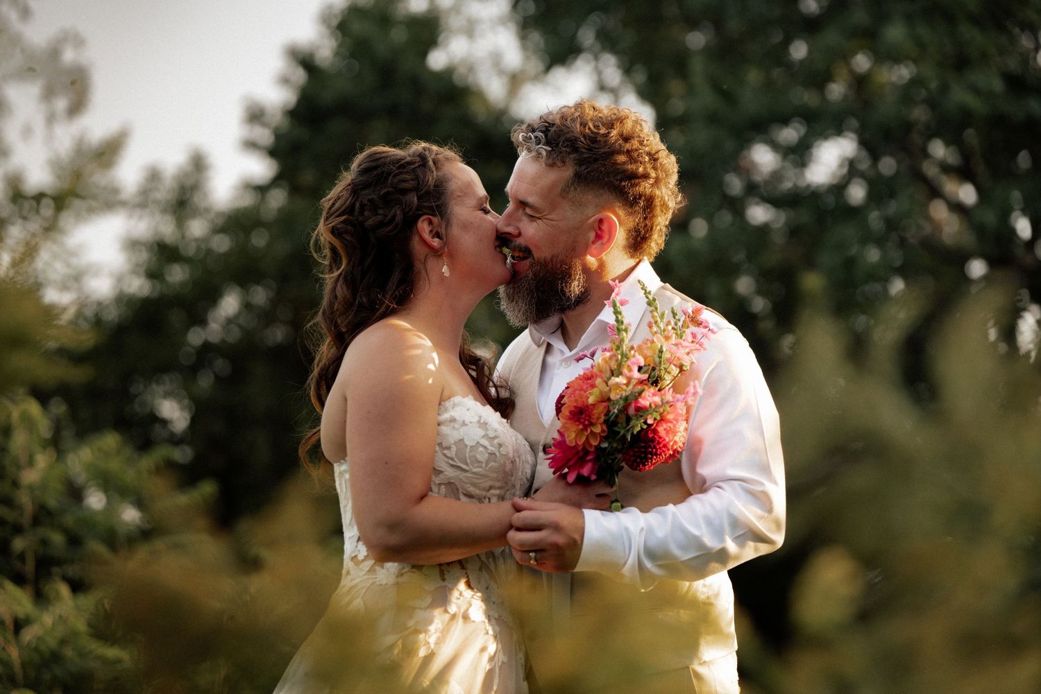 Joyful bride and groom nearly kissing while holding colorful bouquet outdoors.