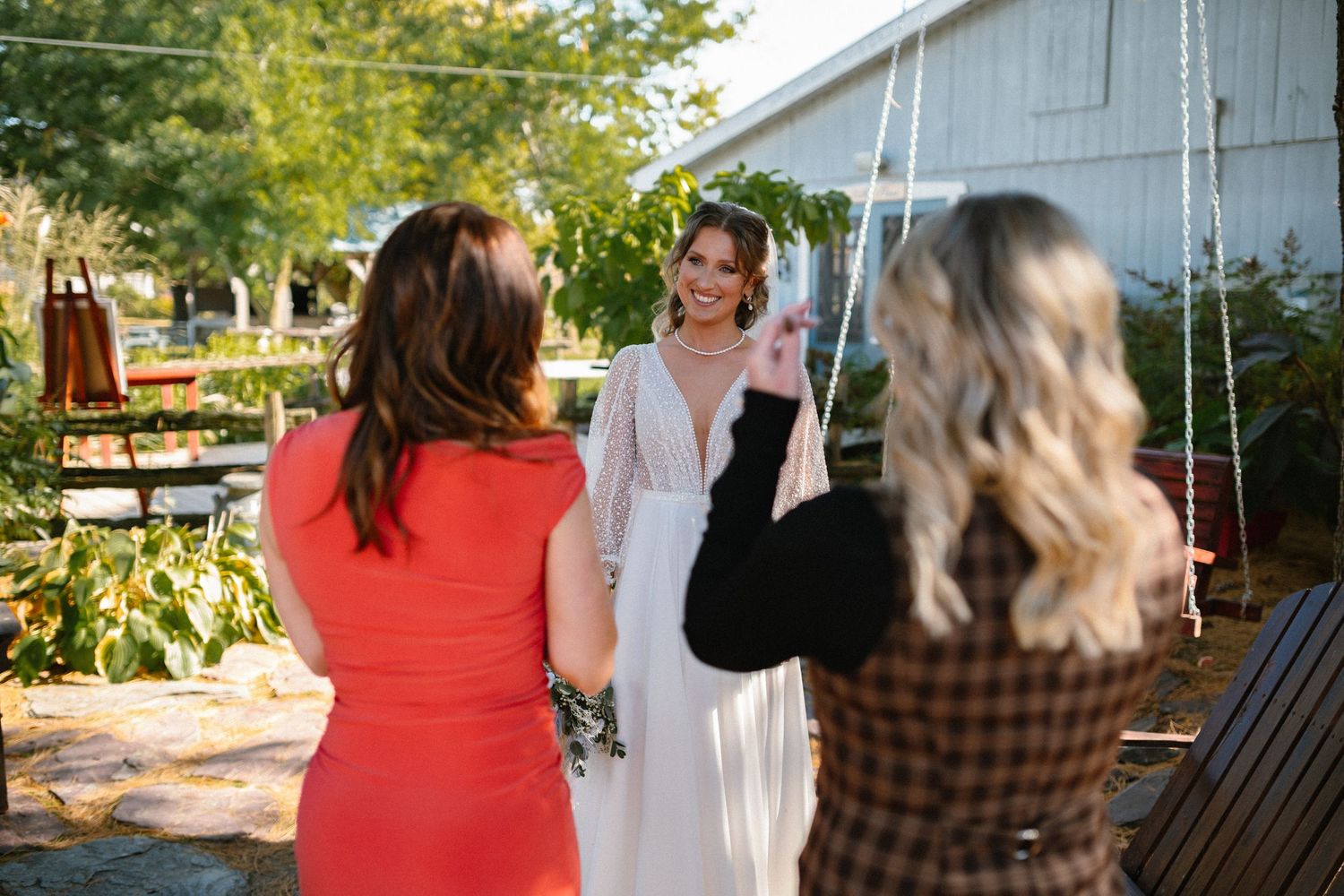 Two women photograph a smiling bride in a white gown outdoors near a swing on a sunny day.