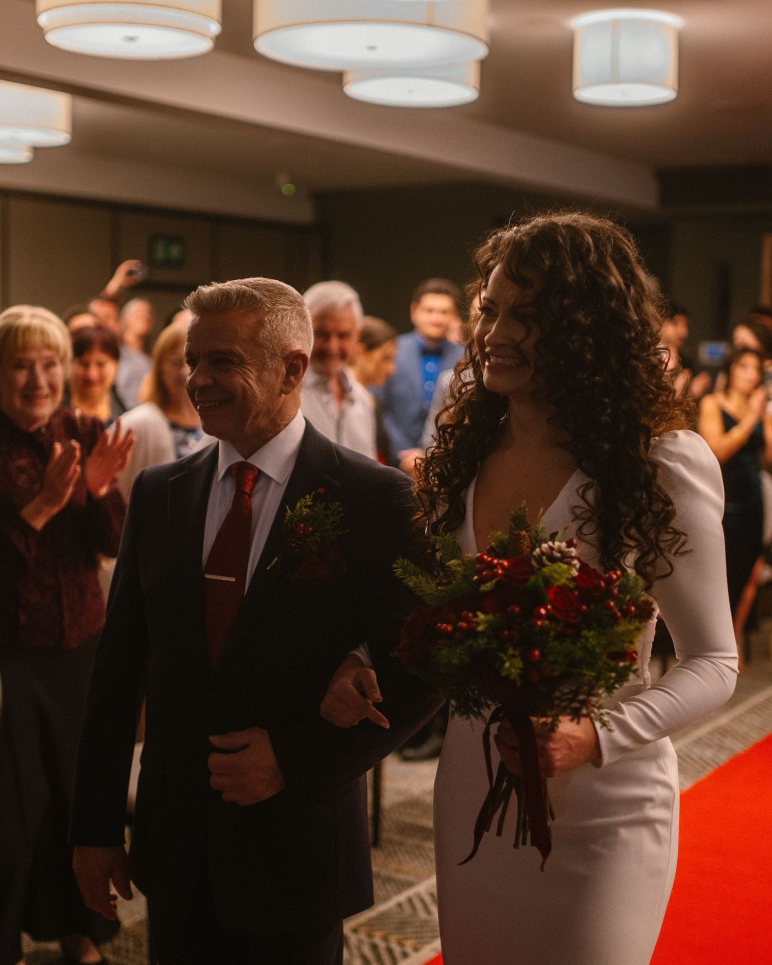 Bride in white dress holding red roses walks down aisle with older man in suit on red carpet at wedding ceremony.