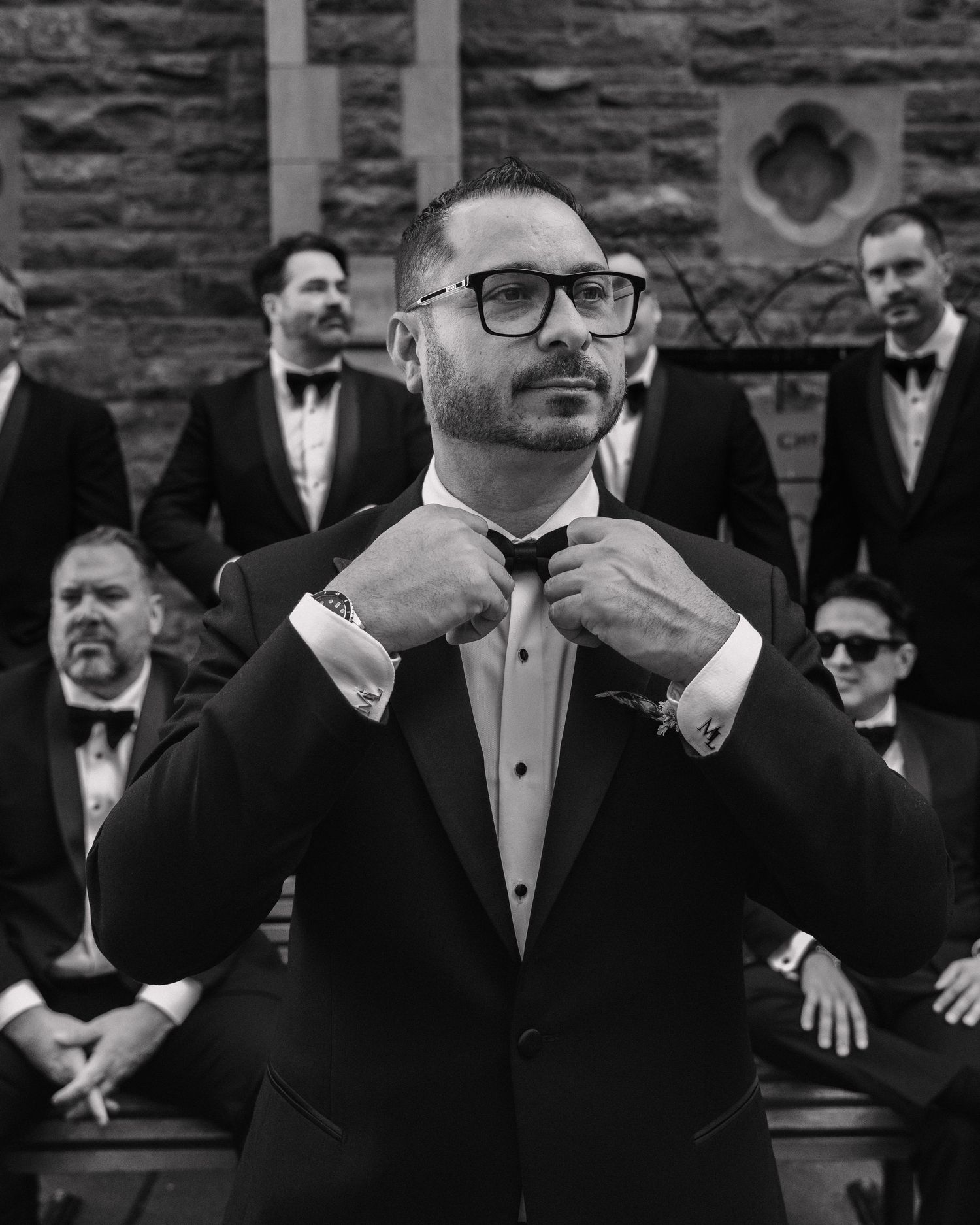 Groom in black tuxedo adjusting bow tie with groomsmen posed behind him in black and white photo.