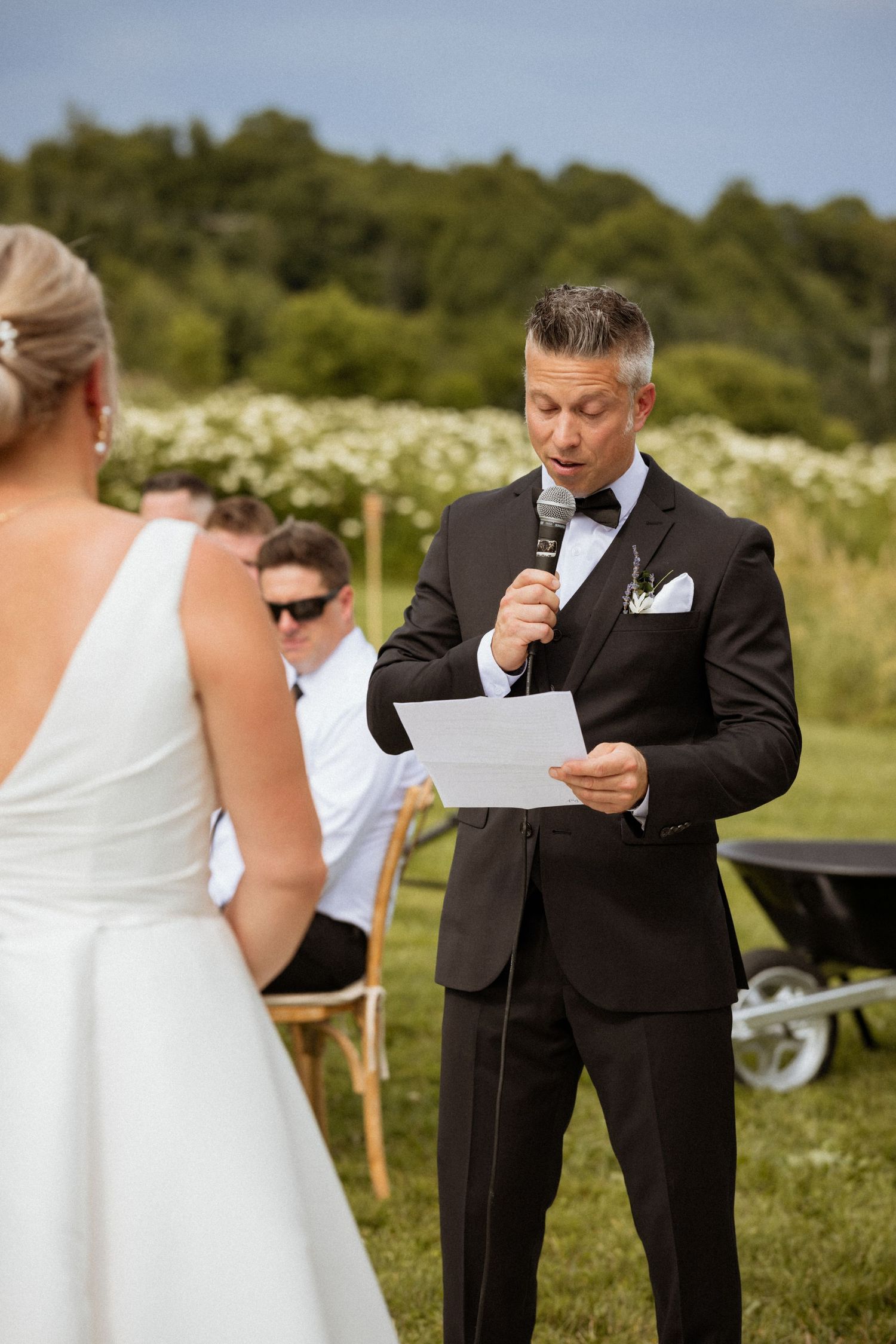 Groom reads vows from paper while holding microphone at outdoor wedding ceremony with bride in white dress.