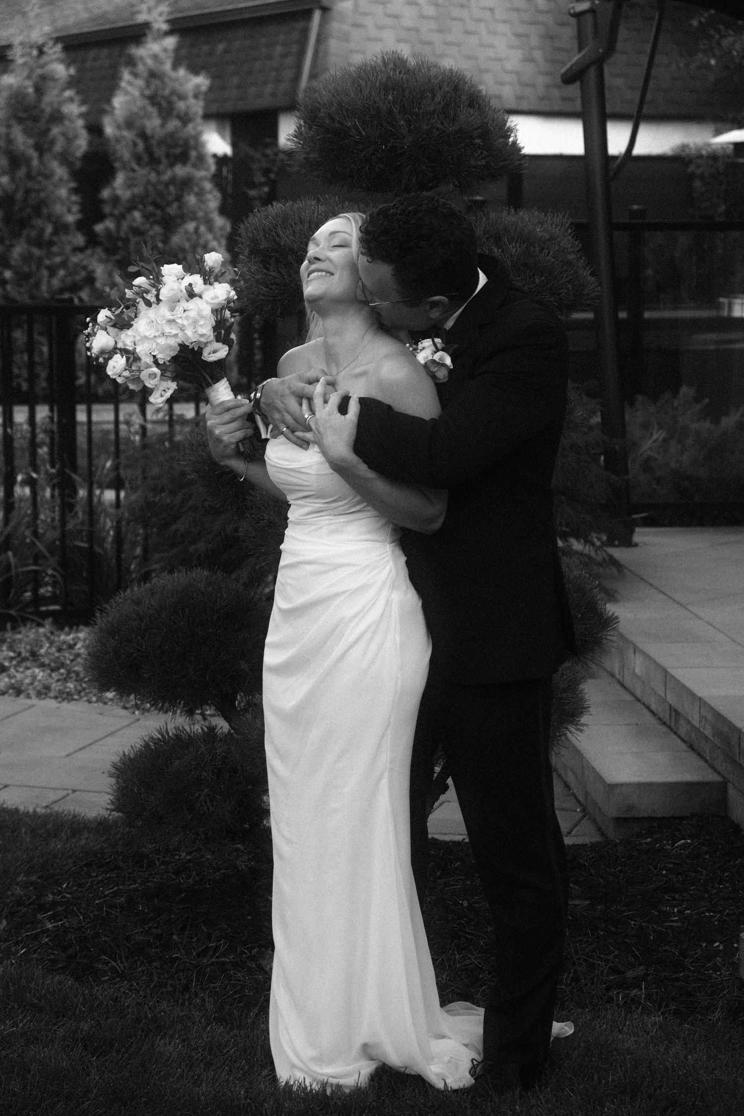 Black and white photo of groom embracing bride from behind outdoors, bride smiling and holding white bouquet.