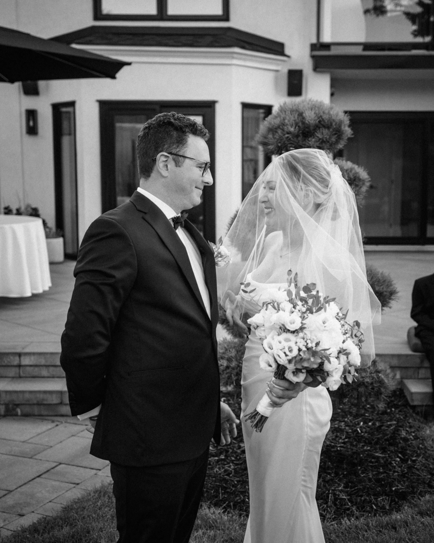 Groom in tuxedo and bride in white gown with veil share a tender moment in black and white photo.