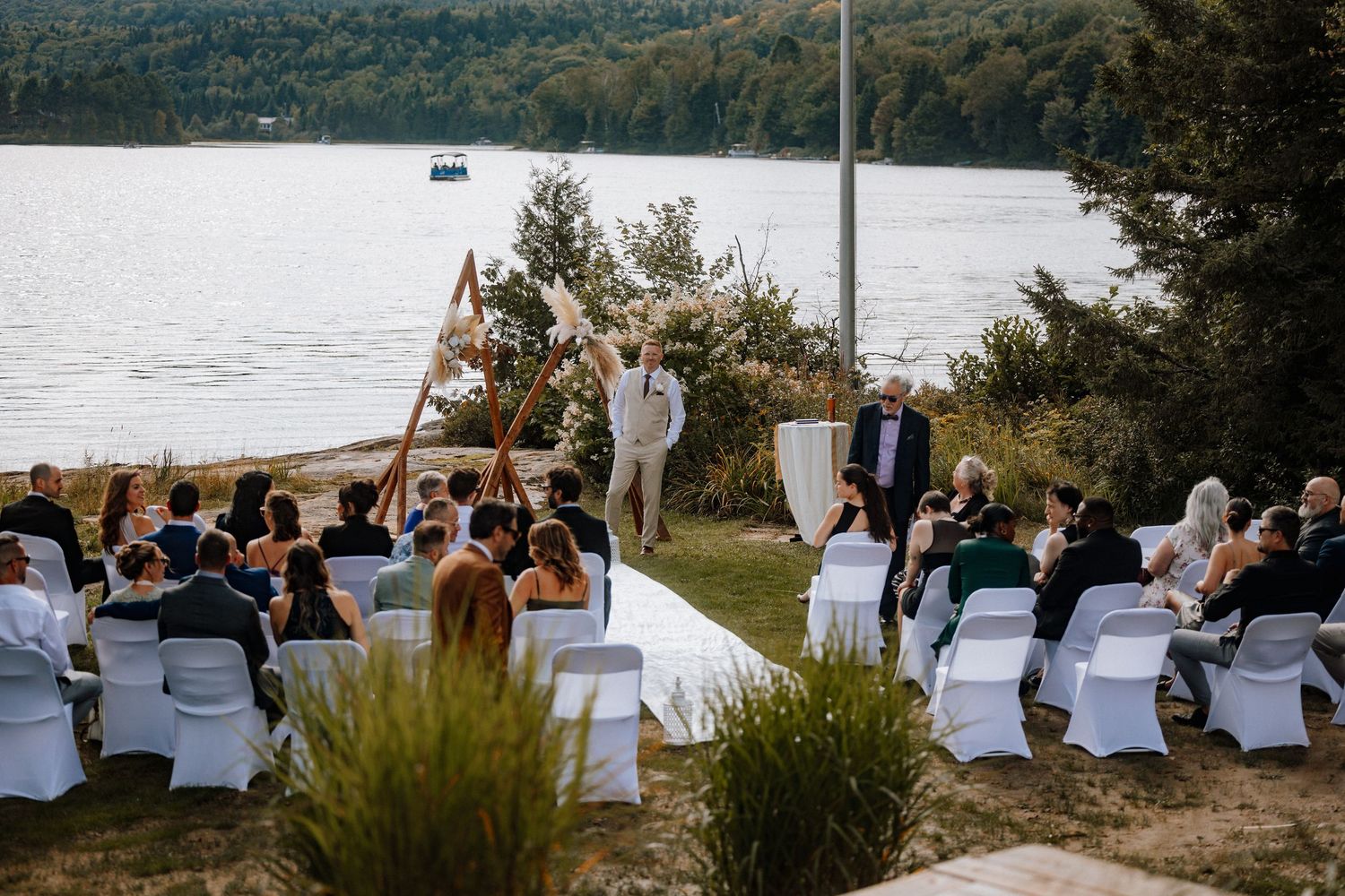 Outdoor lakeside wedding ceremony with white chairs, floral arch, and lush forest backdrop.