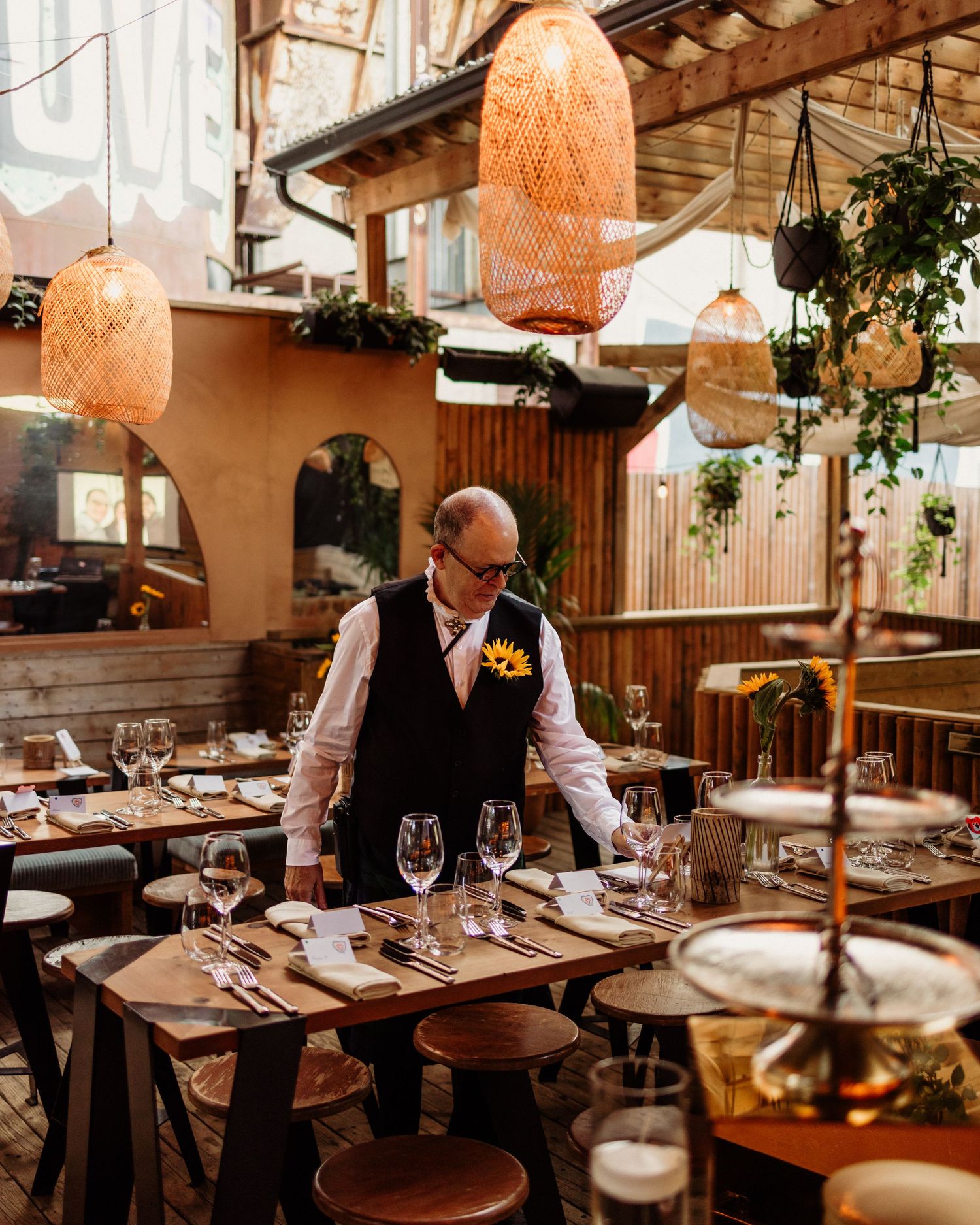 Waiter setting an elegant restaurant table with wine glasses under warm wicker pendant lights.