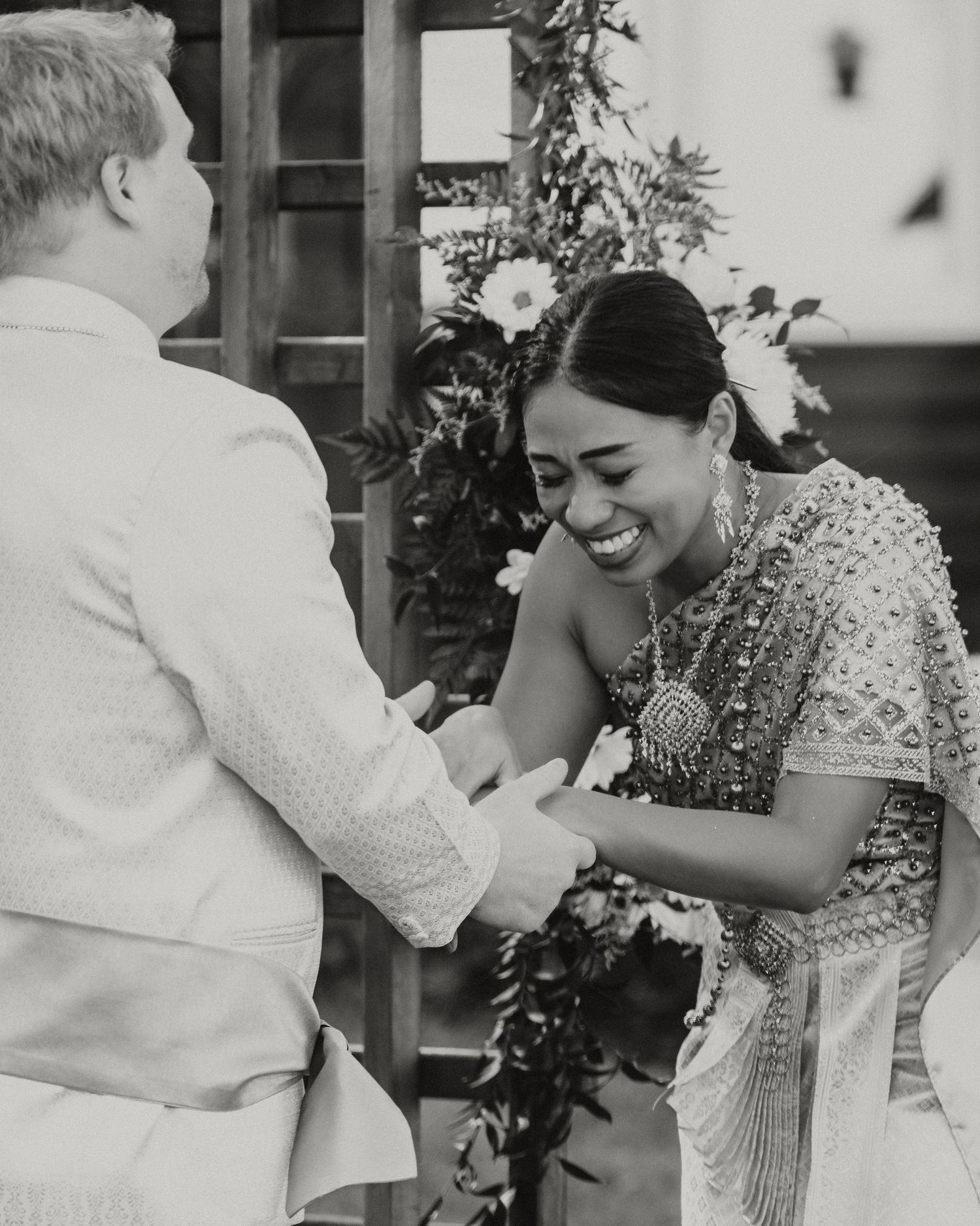 Bride in traditional Indian attire smiles joyfully while exchanging vows with groom in white suit at wedding ceremony.