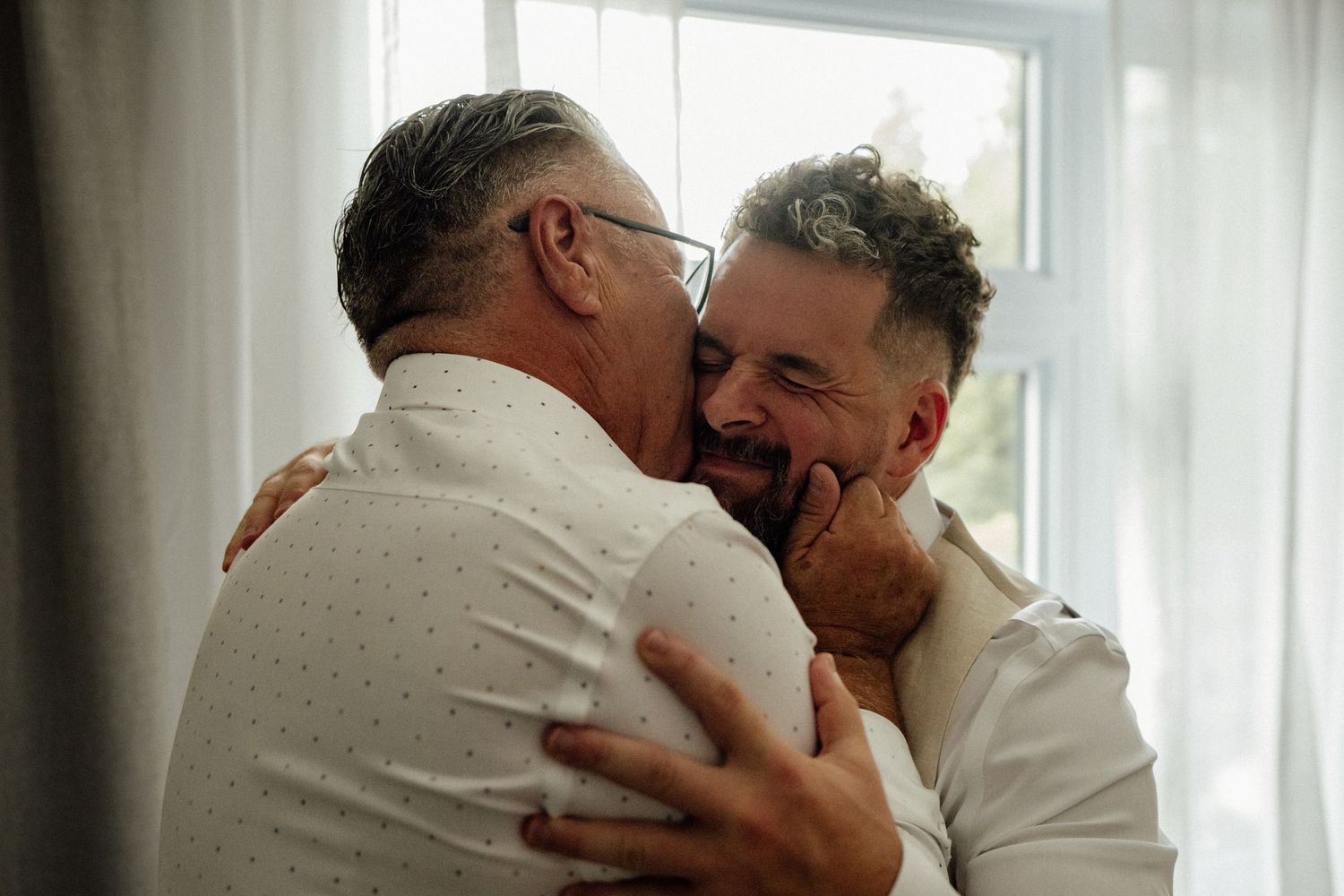 Two older men share a warm, joyful embrace and smile together near a bright window.