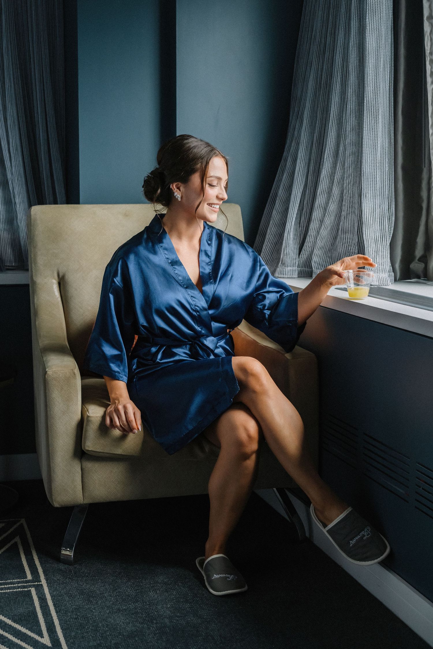 Bride in blue satin robe sitting in armchair by window holding champagne glass on wedding morning.