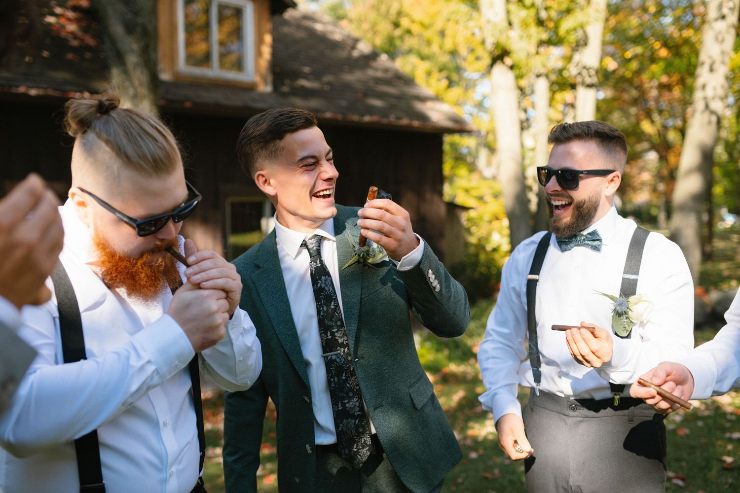 Laughing groomsmen in sunglasses and suspenders celebrating with cigars outside a rustic cabin.