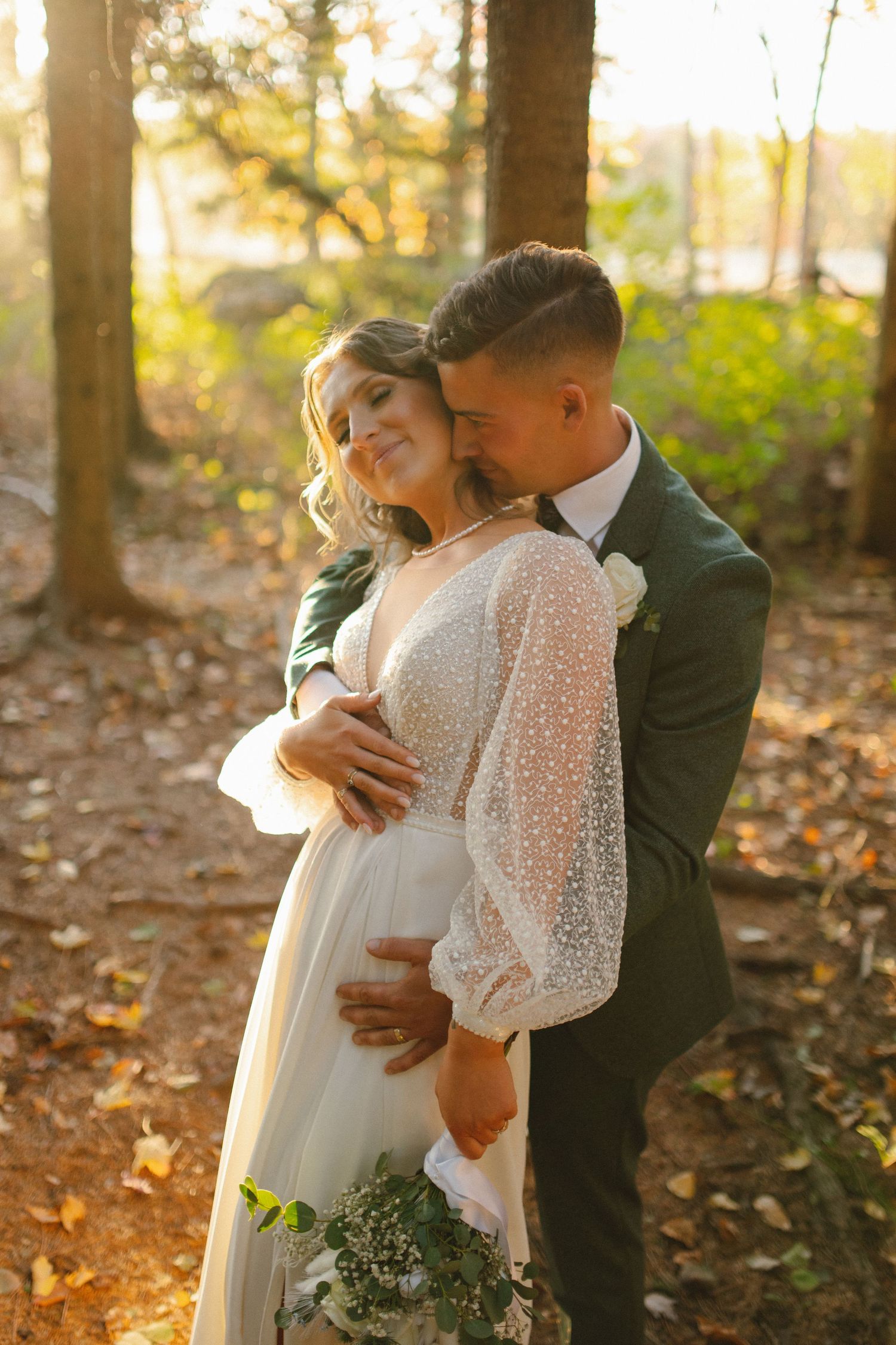 Groom in dark green suit embraces bride in pearl-dotted gown in sunlit autumn forest setting.