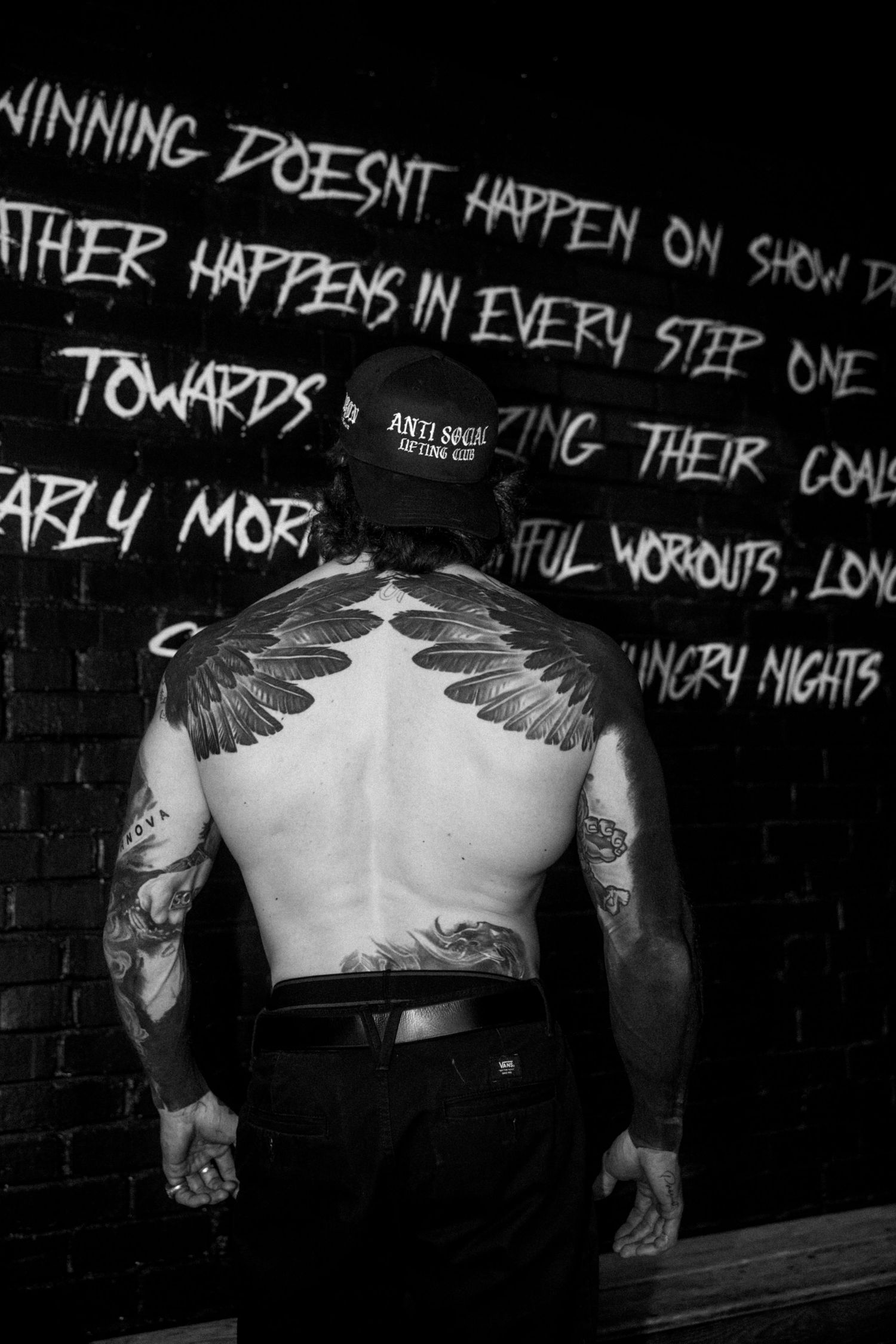 Tattooed man showing large wing back tattoo standing before motivational graffiti wall art.
