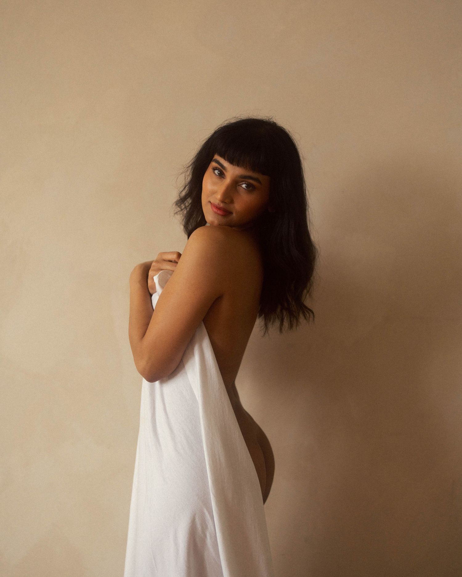 Dark-haired woman in backless white dress looking over shoulder against beige background.