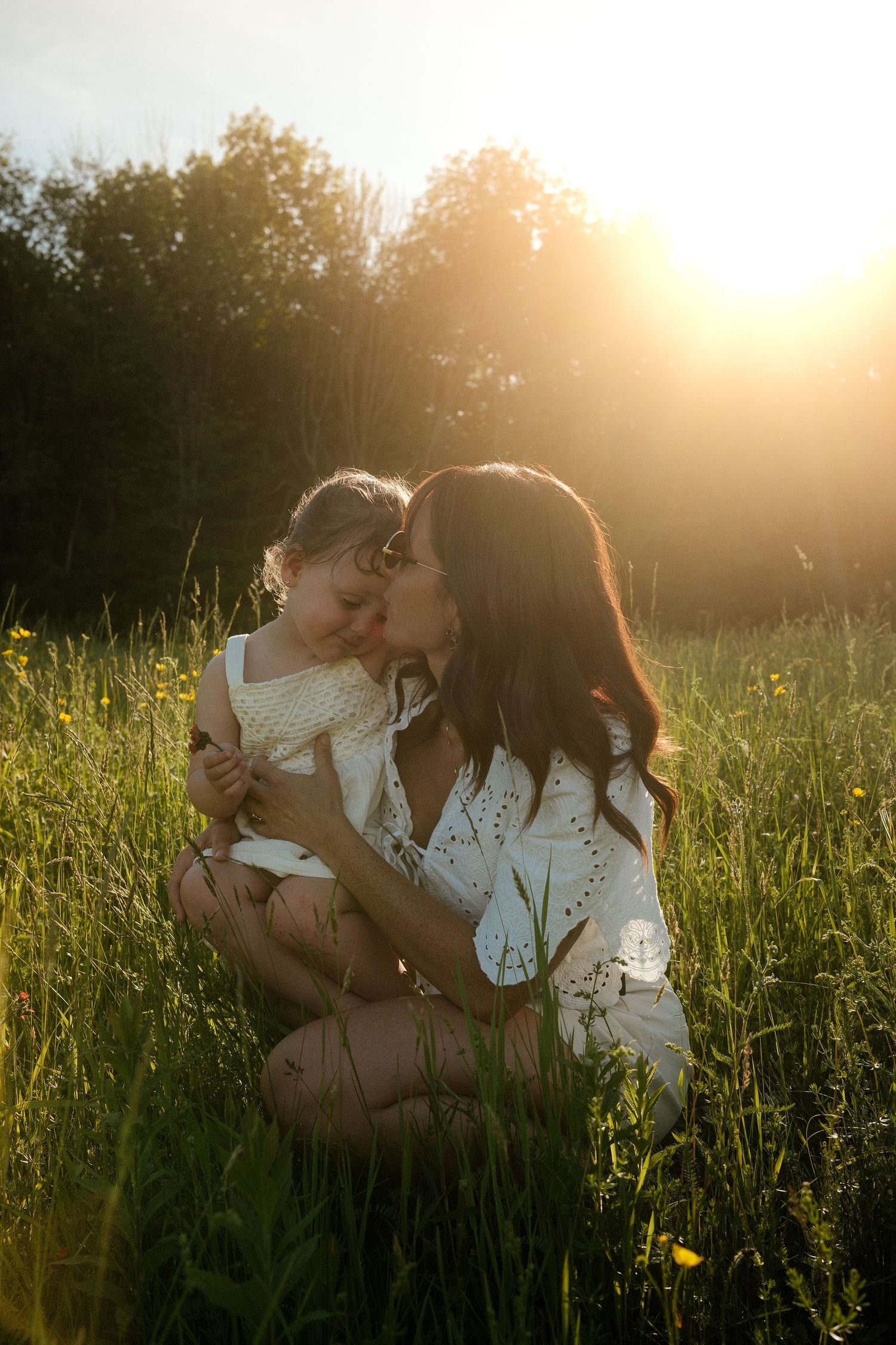 A mother tenderly embraces her young daughter in a sunlit green meadow during golden hour.