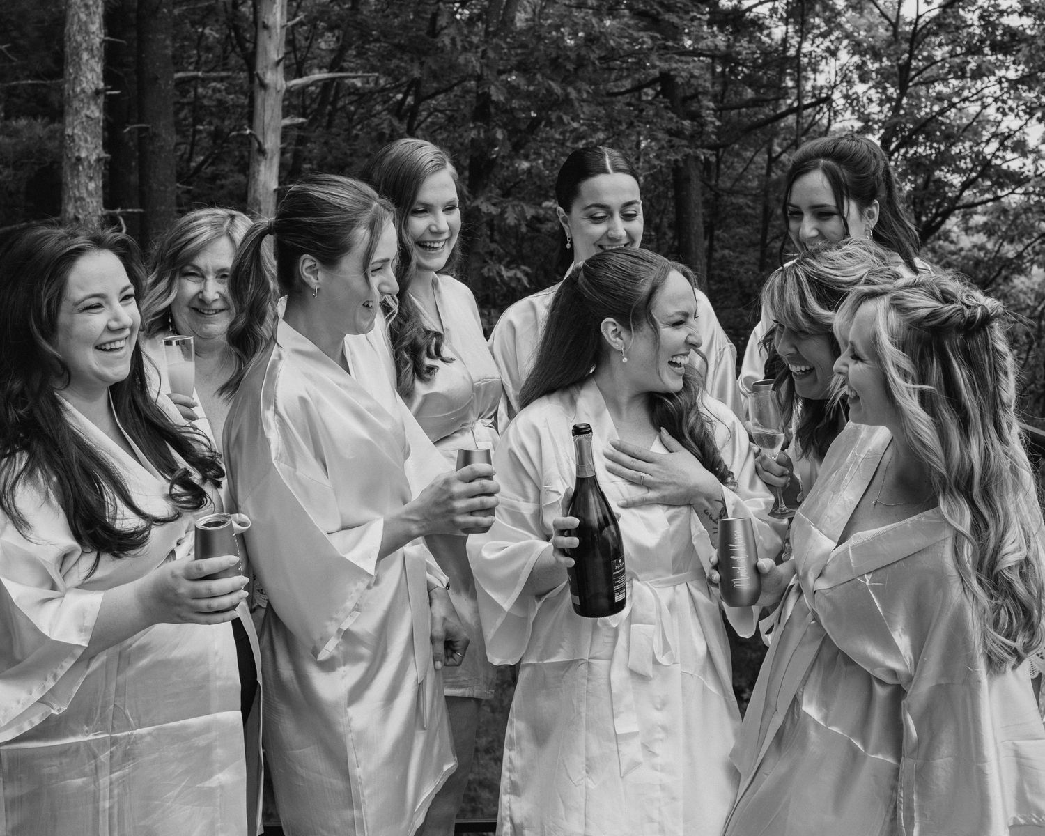 Bridesmaids in white robes celebrate with champagne outdoors in a black and white wedding photo.