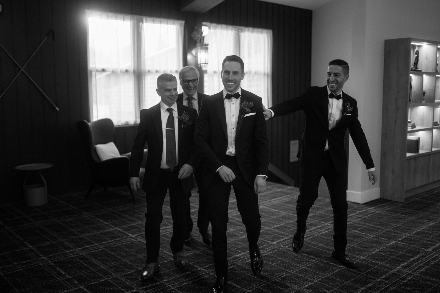 Black and white photo of groom and groomsmen in tuxedos walking confidently through a hotel room.