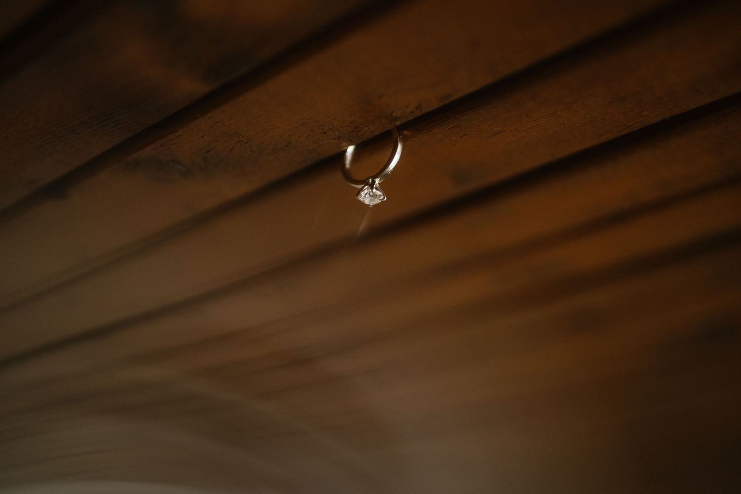 Diamond engagement ring hanging from a wooden ceiling beam in warm light.
