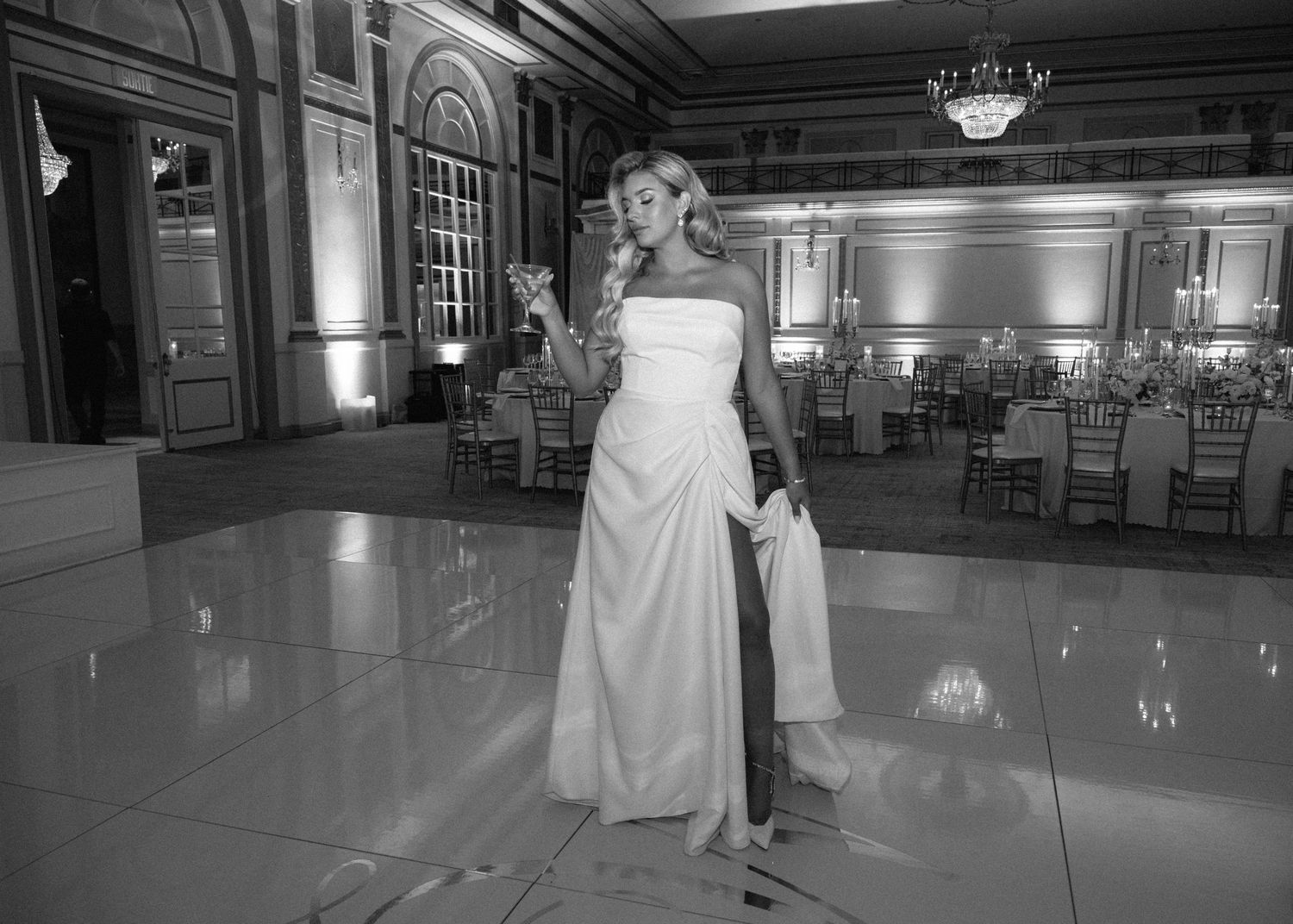 Black and white photo of a bride in a strapless gown posing alone on a ballroom dance floor.
