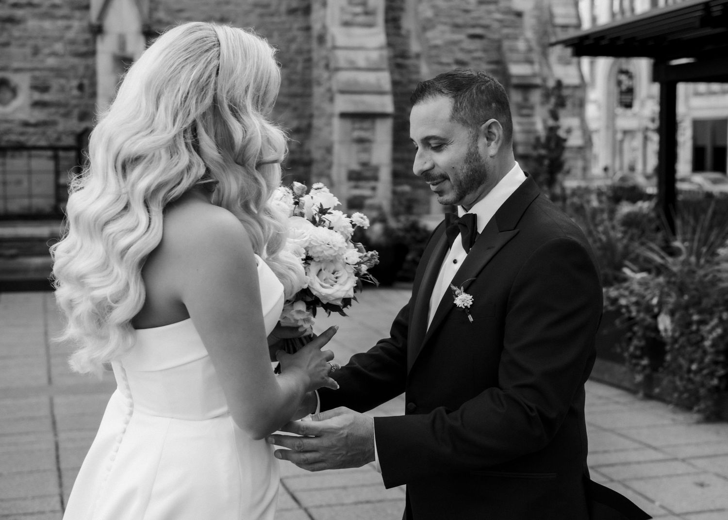 Black and white wedding photo of smiling groom in tuxedo holding bride's bouquet during first look ceremony.