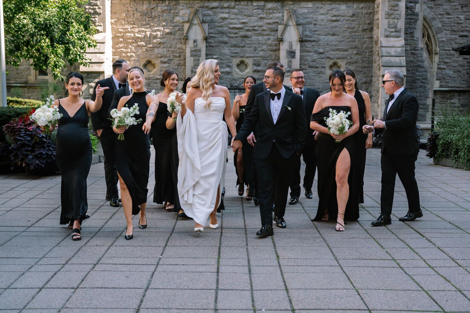 Wedding party walking together outside stone church, bridesmaids in black gowns, groomsmen in tuxedos.