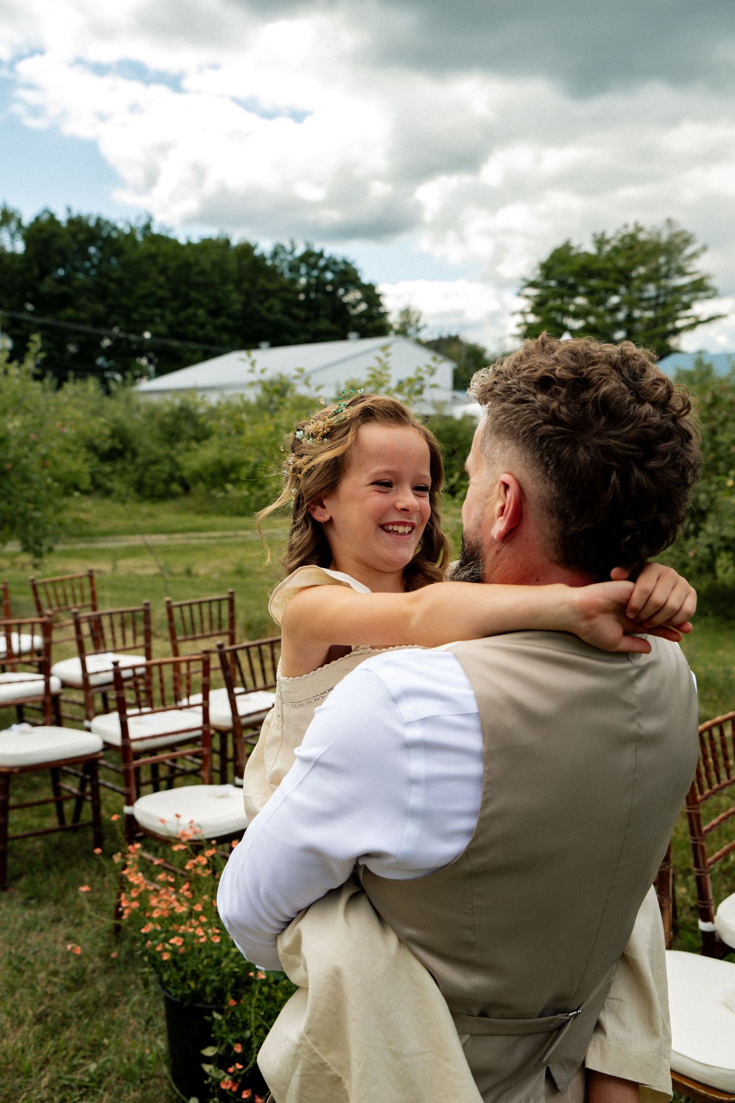 A groom in a tan vest lifts a smiling flower girl at an outdoor wedding ceremony with garden chairs.