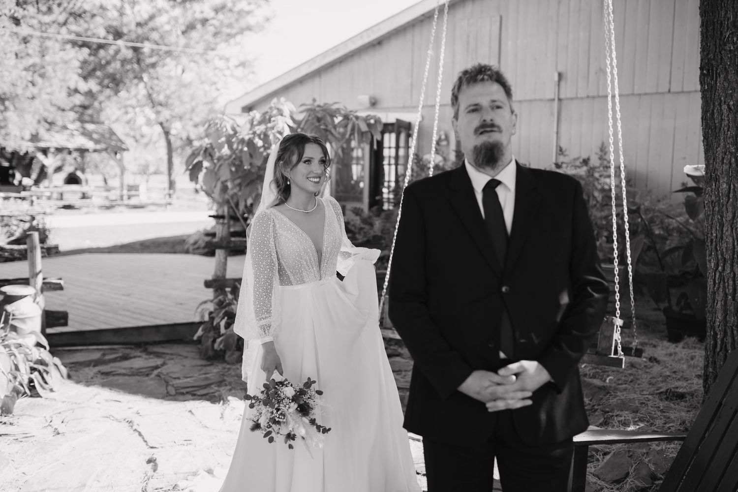 Black and white photo of a bride holding bouquet and groom in suit standing together outdoors at a rustic wedding venue.
