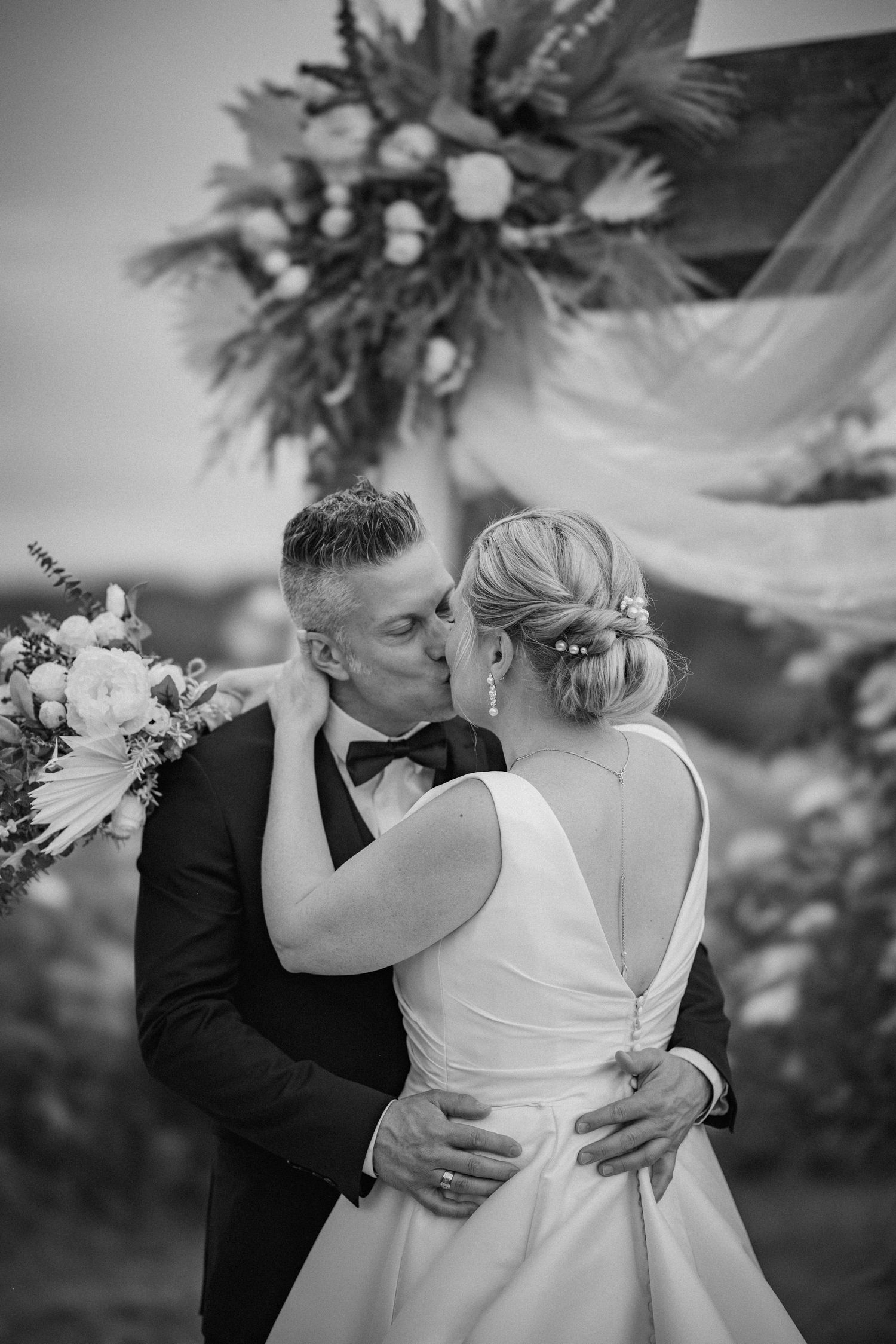 Black and white photo of a couple sharing their first kiss at an outdoor wedding ceremony beneath a floral arch.