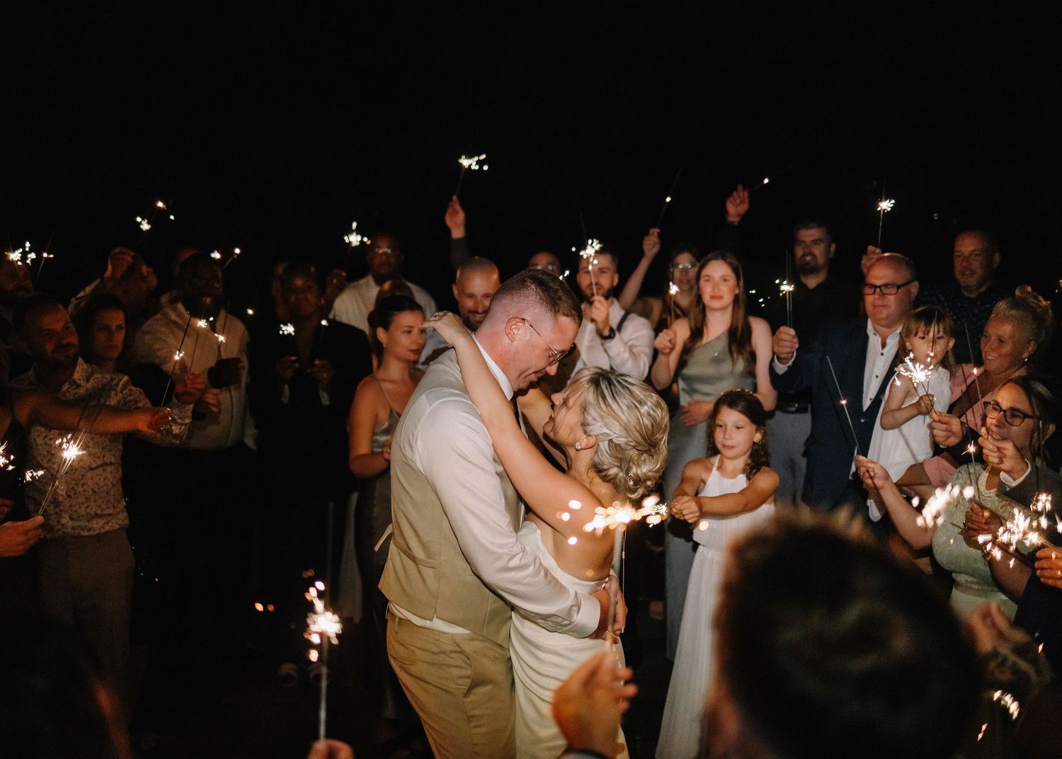 Newlyweds share romantic kiss surrounded by cheering guests holding sparklers at night wedding sendoff.