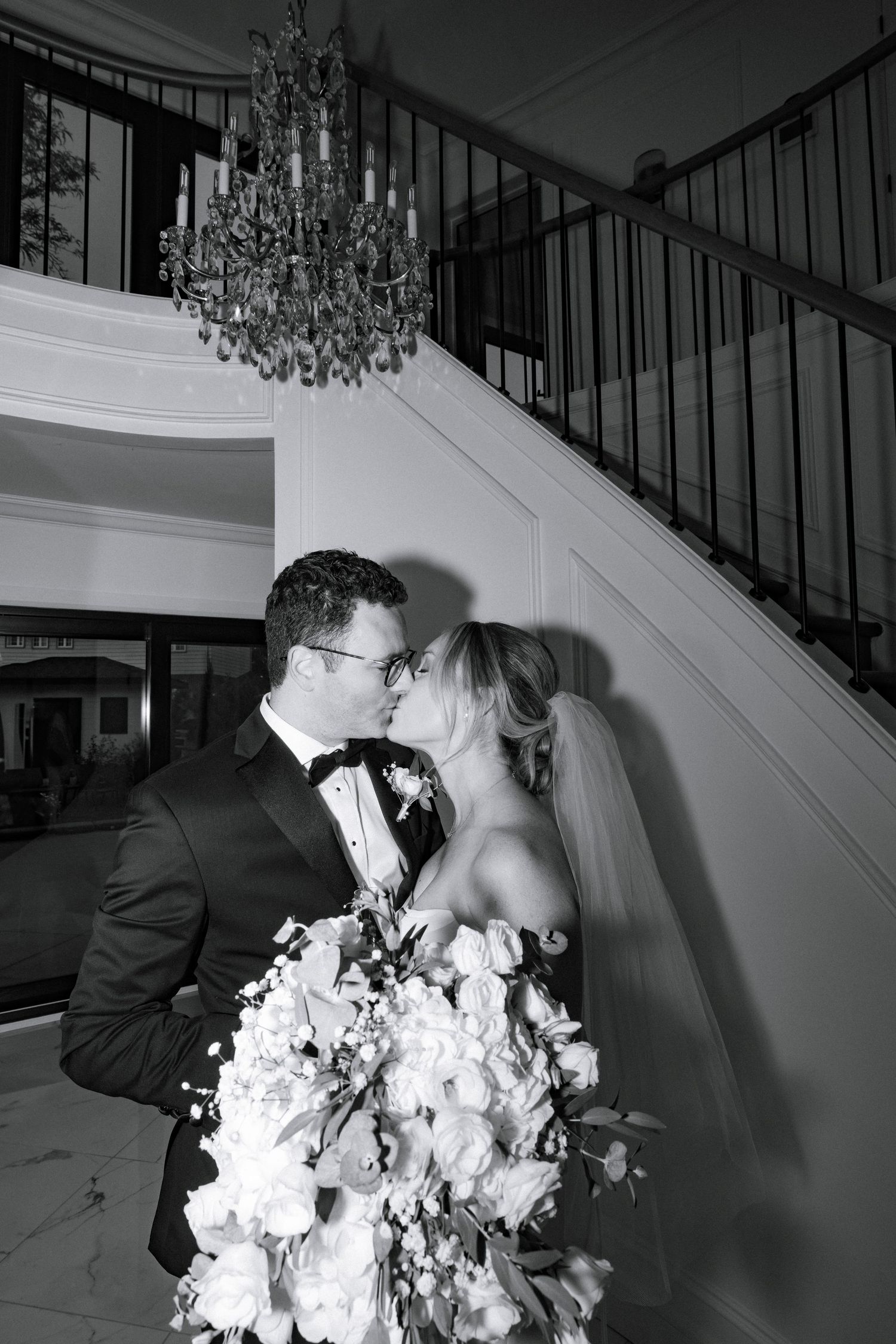 Black and white photo of a bride and groom kissing near a grand staircase with chandelier.