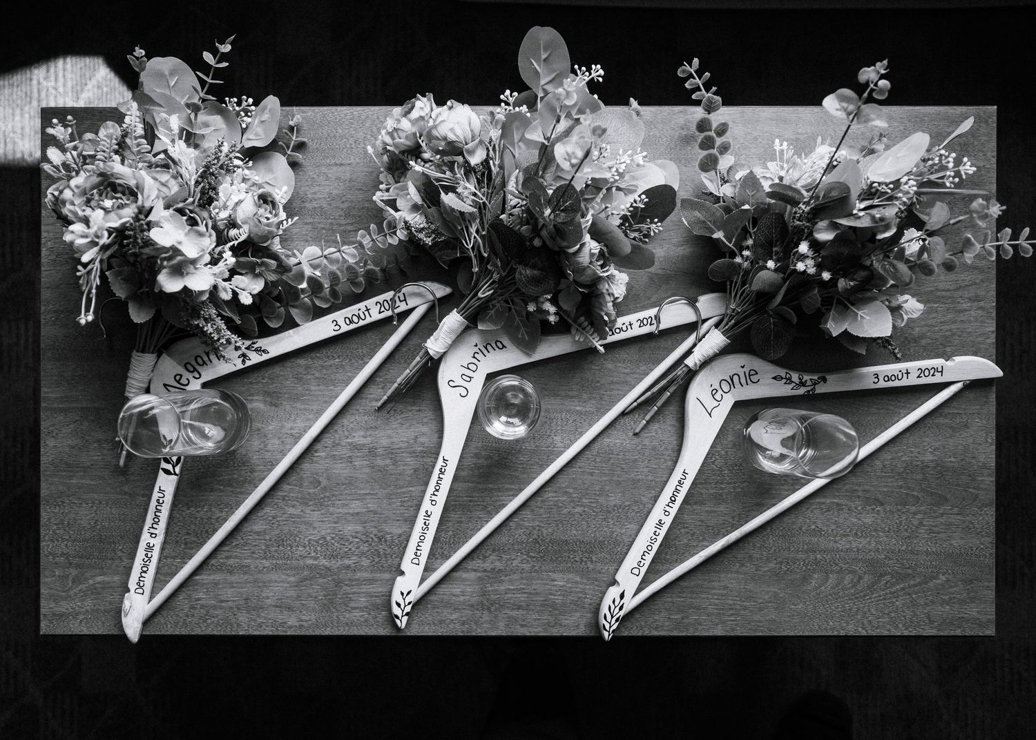 Black and white photo of personalized wooden wedding hangers with bridal bouquets on a wooden table.