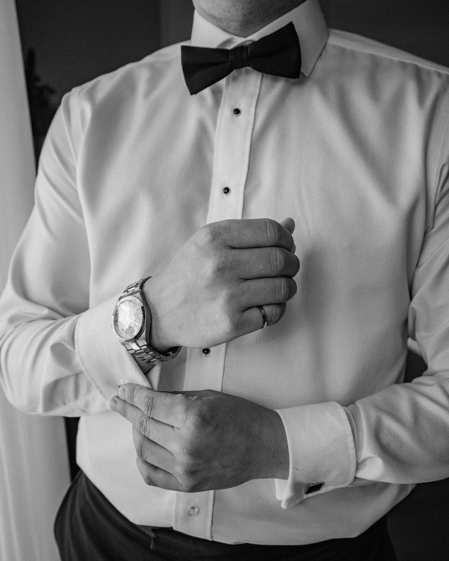 Groom adjusting cufflinks wearing white tuxedo shirt with bow tie and silver watch before wedding ceremony.