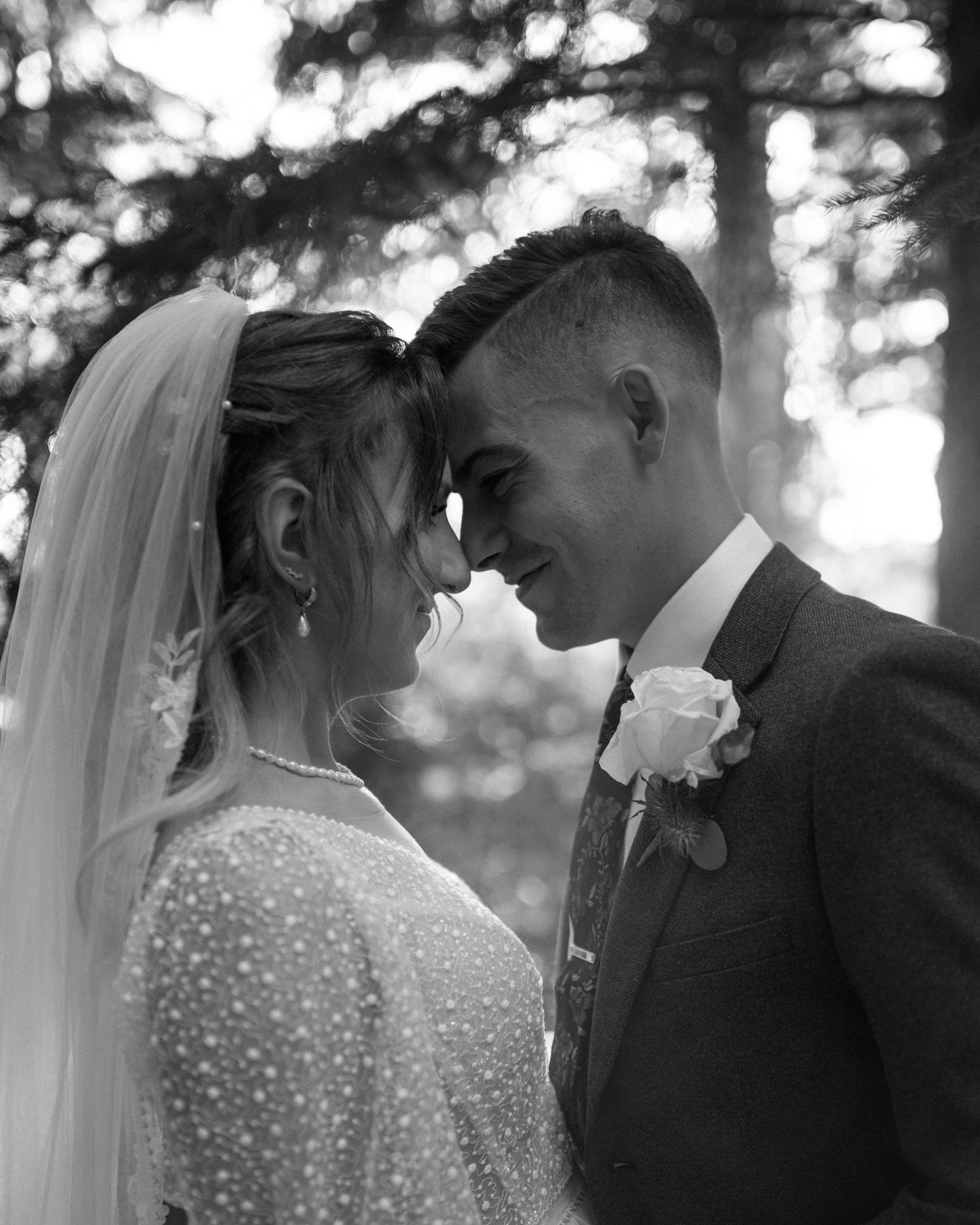 Black and white close-up of bride and groom smiling forehead to forehead in forest.