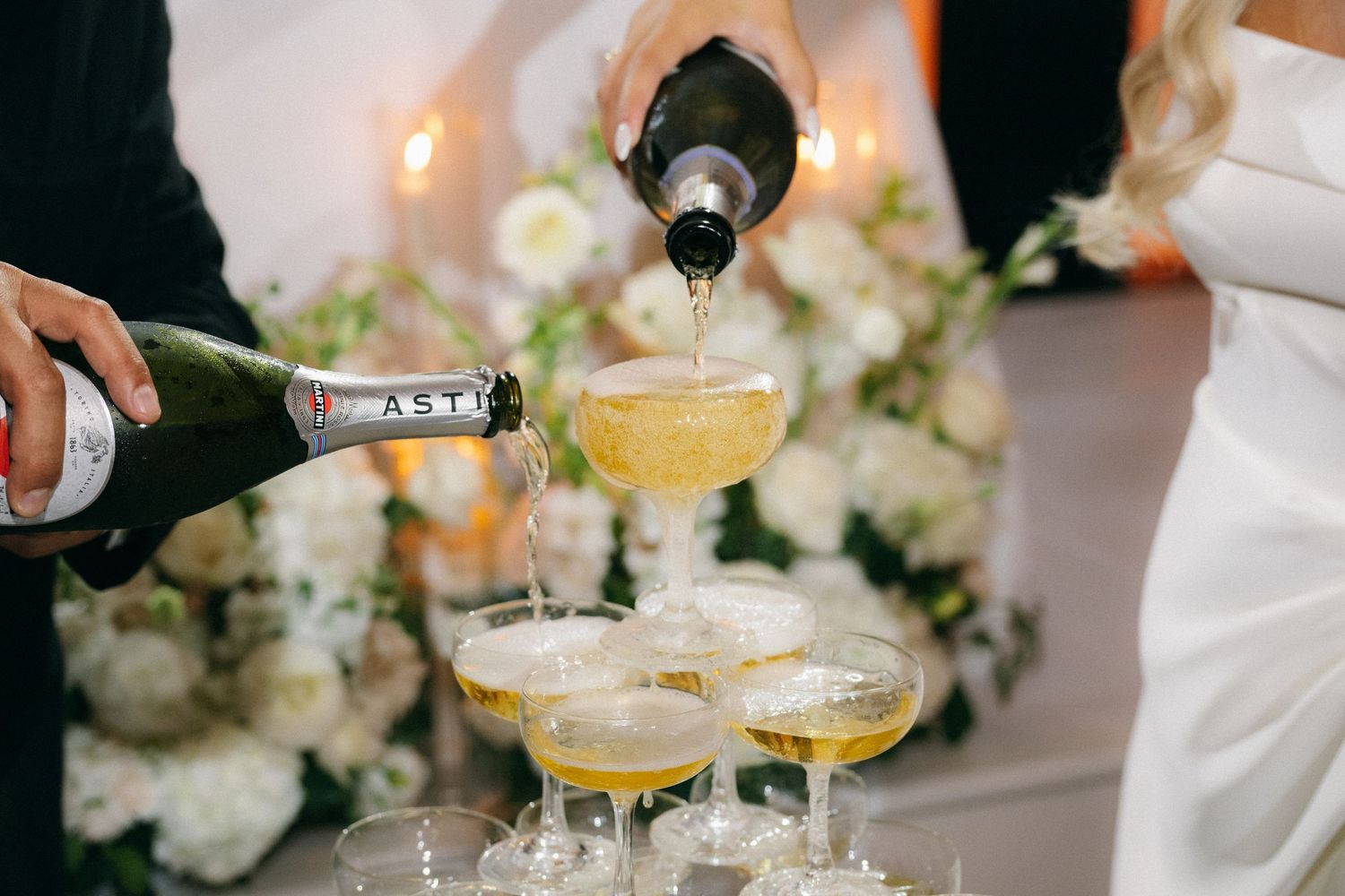 Champagne tower being filled with sparkling wine surrounded by white floral arrangements and candles.