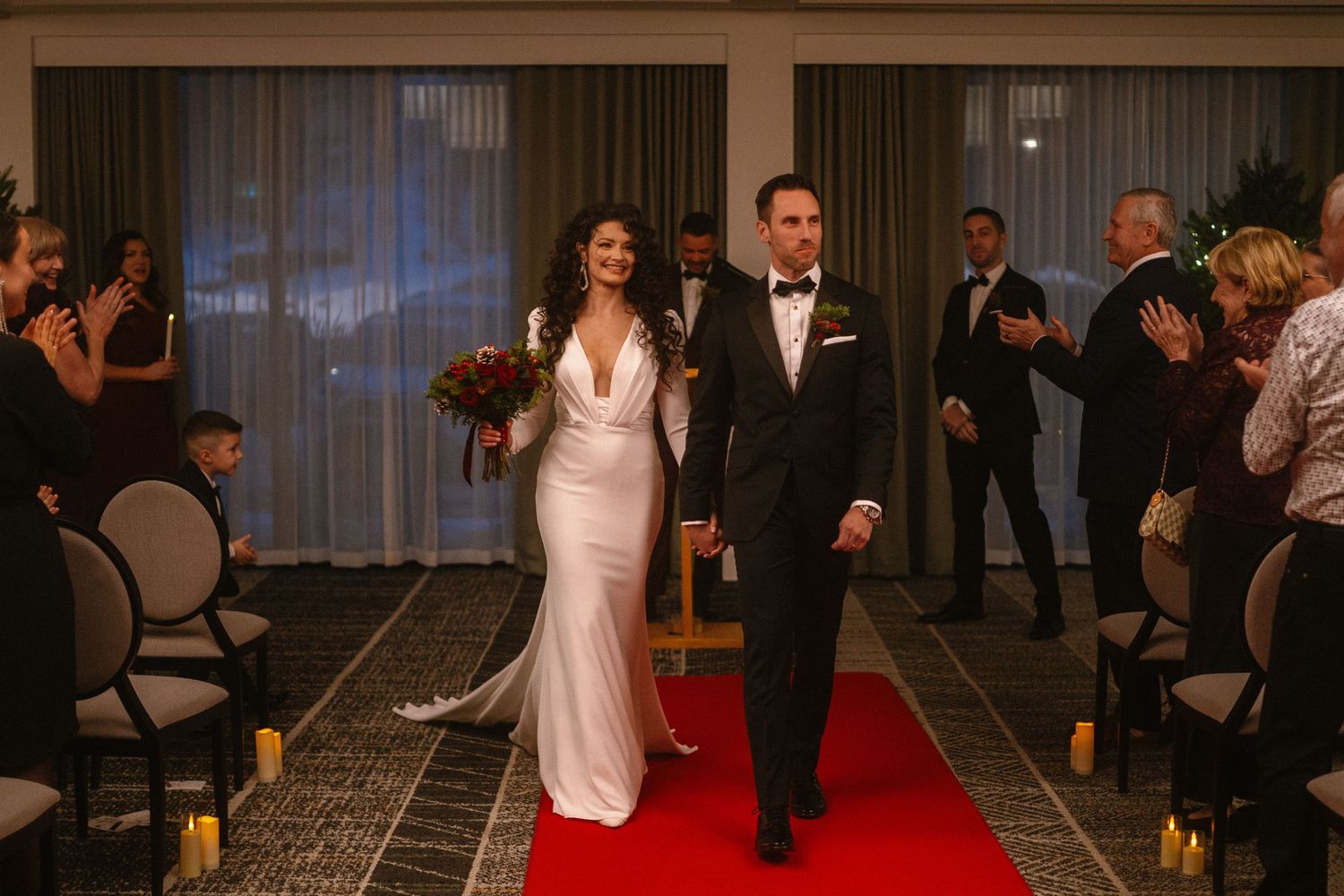 Bride in white gown and groom in black tuxedo walk down red carpet aisle at elegant indoor wedding ceremony.