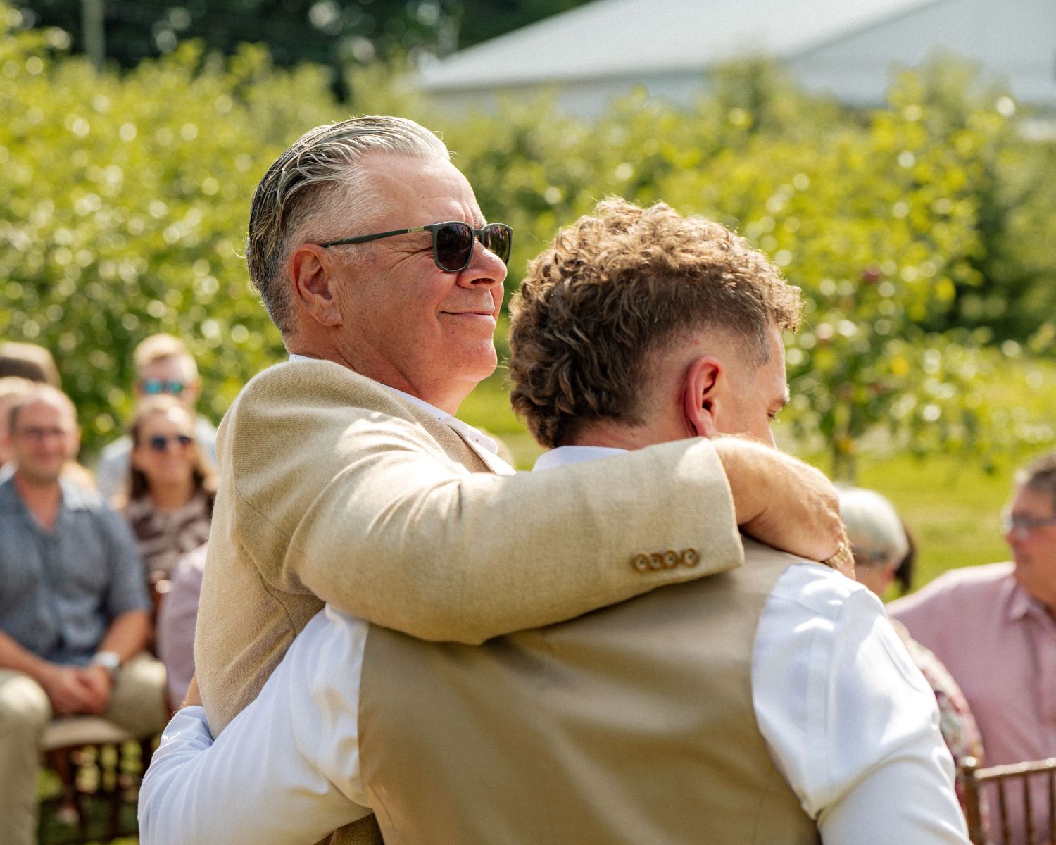 Two men in tan suits share a warm hug at an outdoor wedding ceremony on a sunny day.