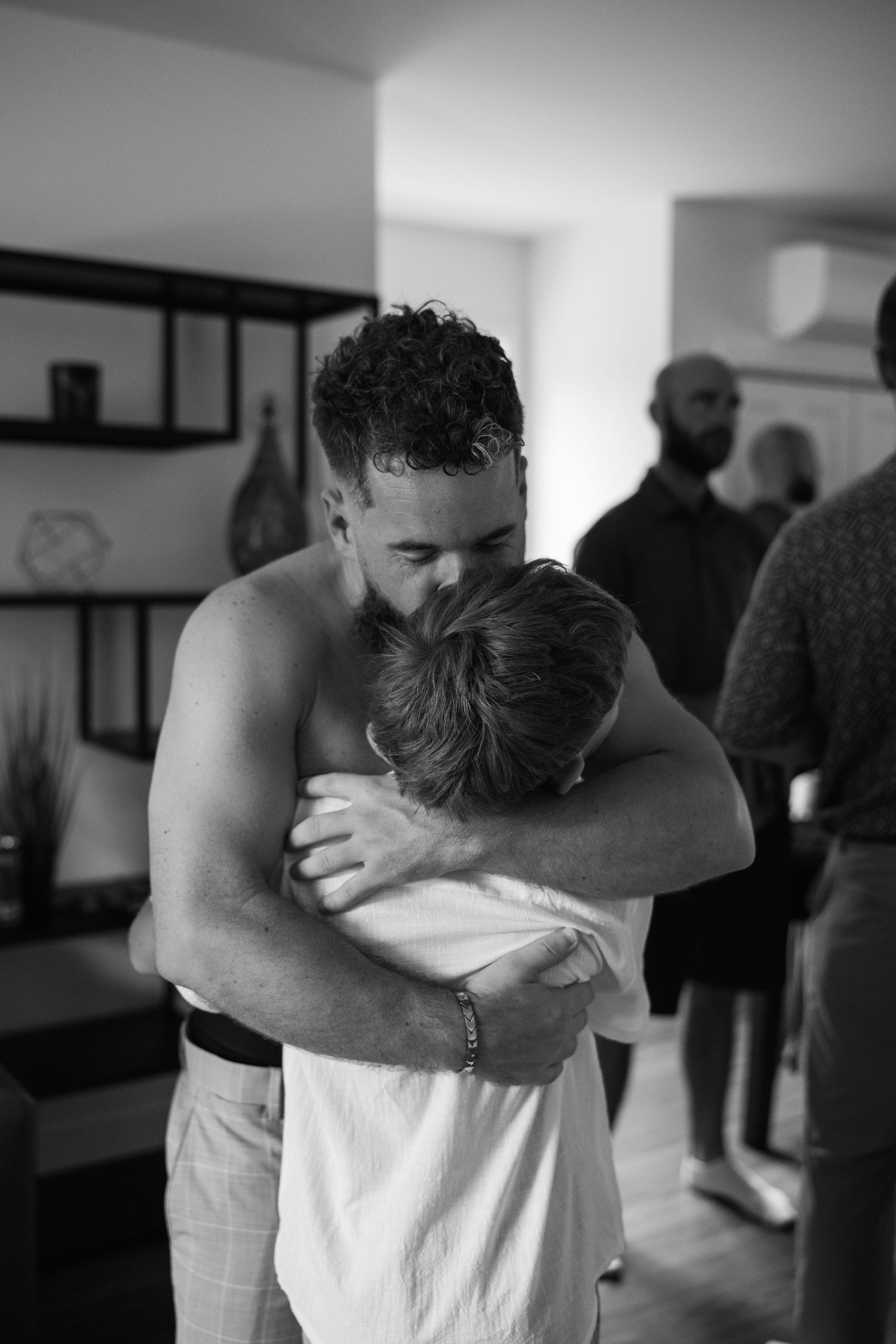 Shirtless man shares a heartfelt embrace with a young boy in a black and white candid indoor photo.