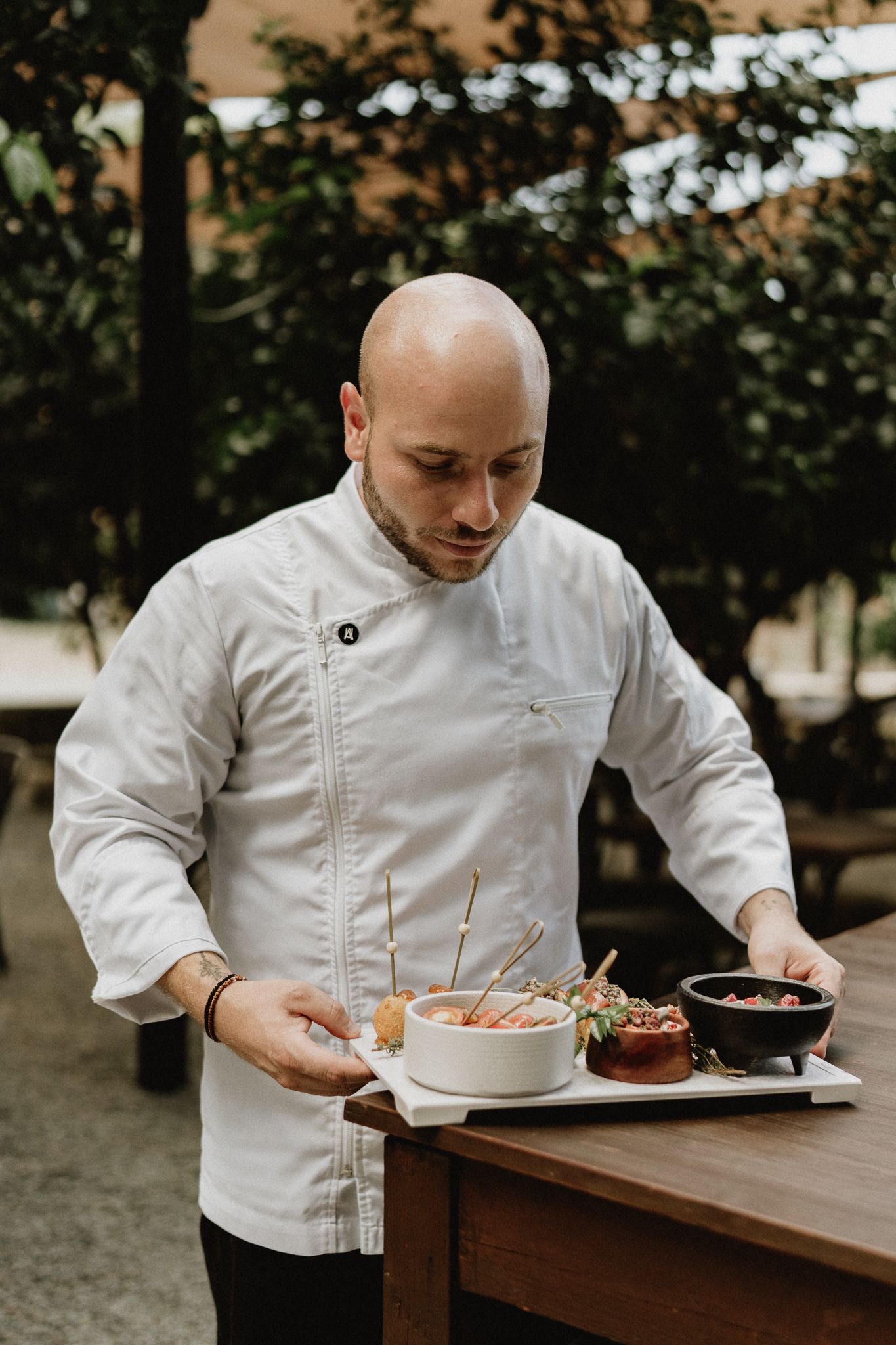 A bald chef in white uniform carefully presents a gourmet dish on a wooden table in an outdoor restaurant setting.