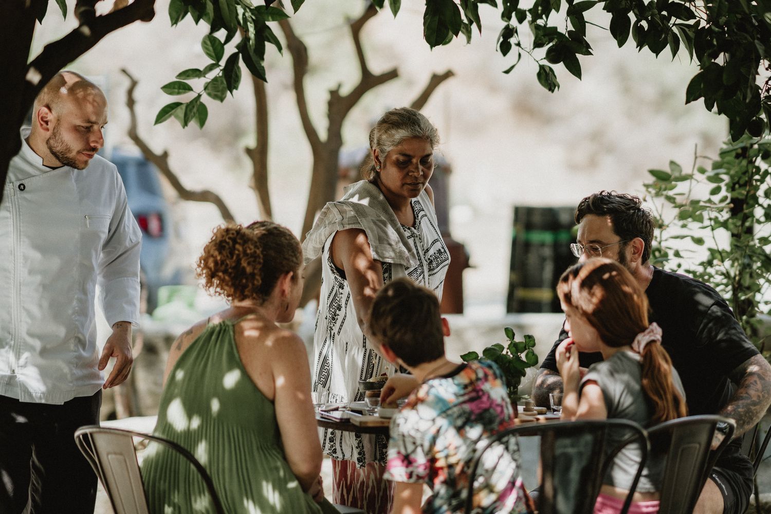 Family and friends gathering outdoors under trees enjoying a meal together at a casual garden dinner.