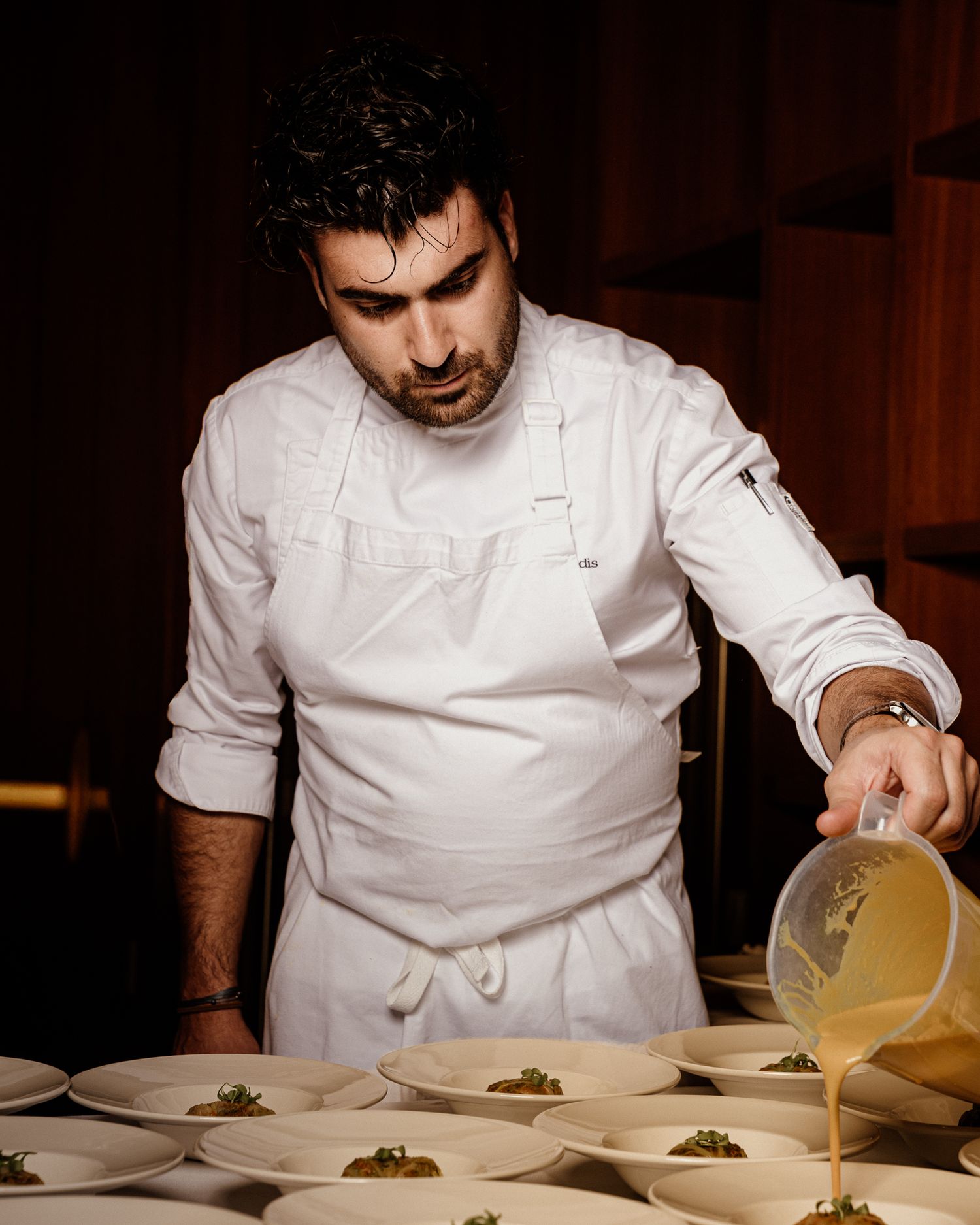 Chef in white coat pouring broth over plated dishes in a fine dining restaurant kitchen.