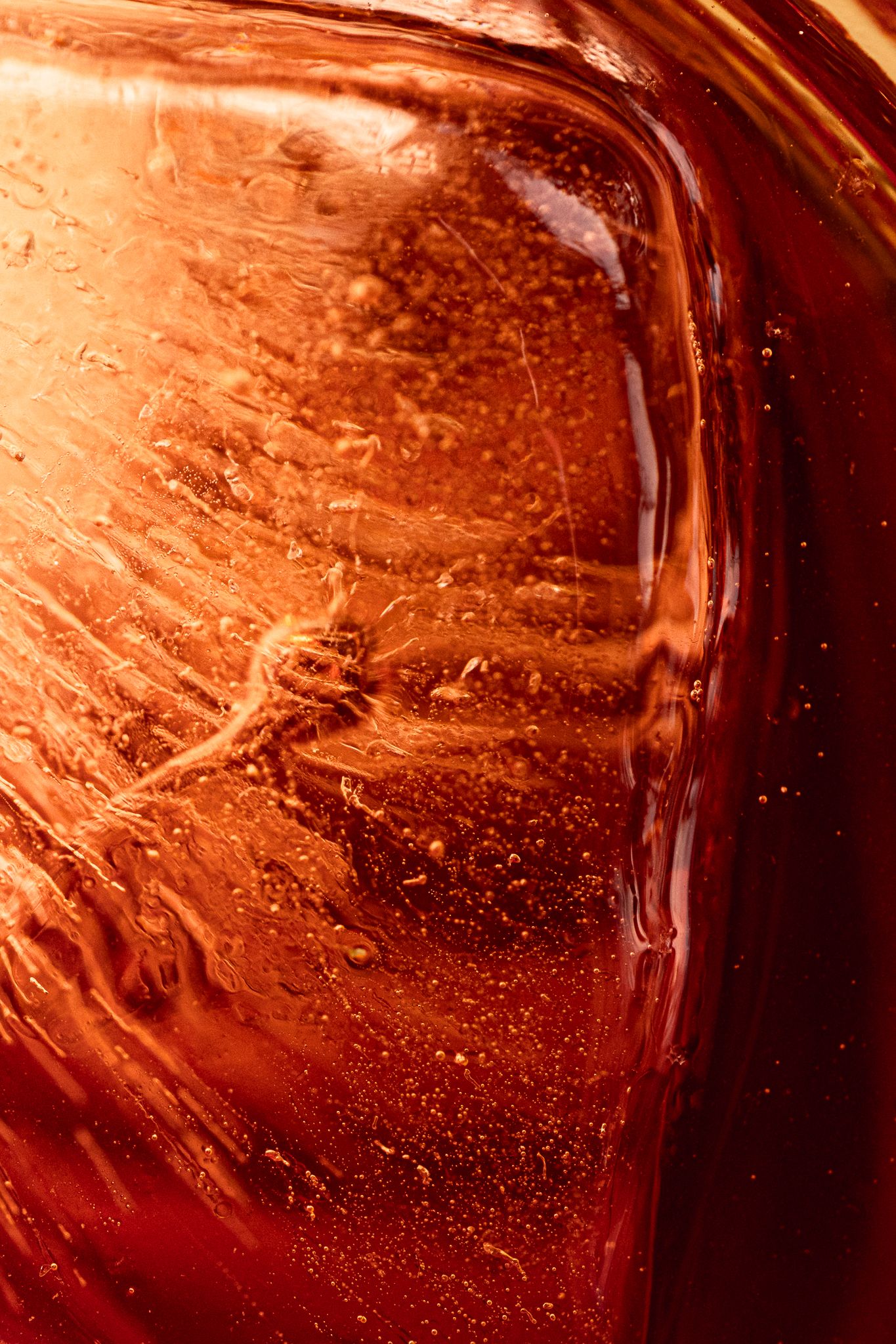 Close-up macro shot of rich amber-red liquid inside a glass jar with bubbles and swirling patterns.
