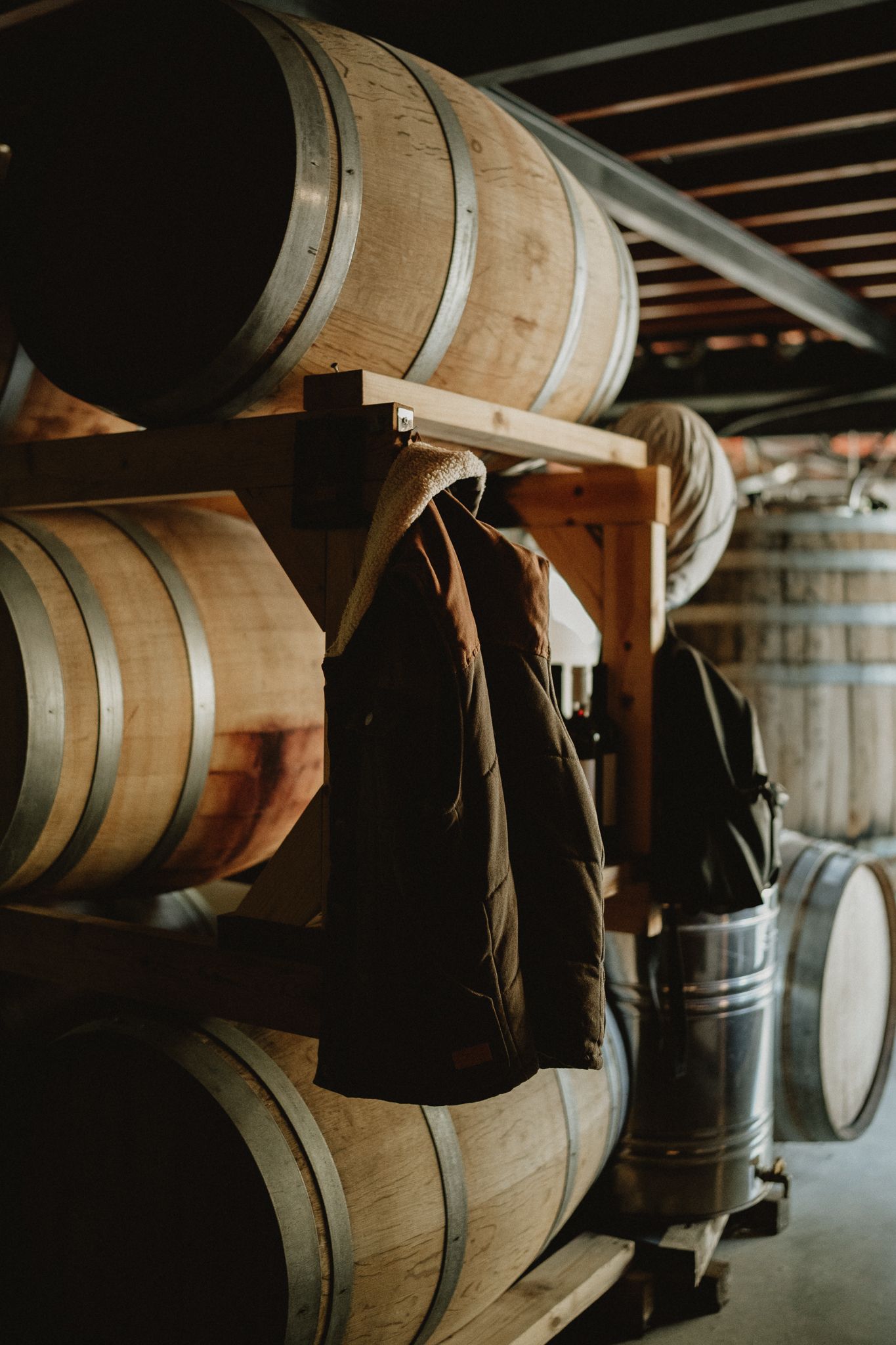 Wooden wine barrels stacked on racks in a cellar with a dark jacket hanging on the barrel rack.